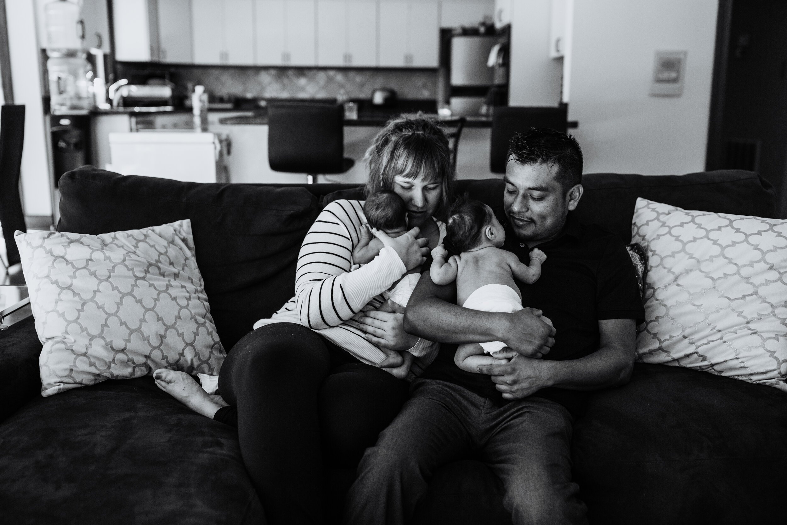 new parents hold twin baby close while sitting on couch at home 
