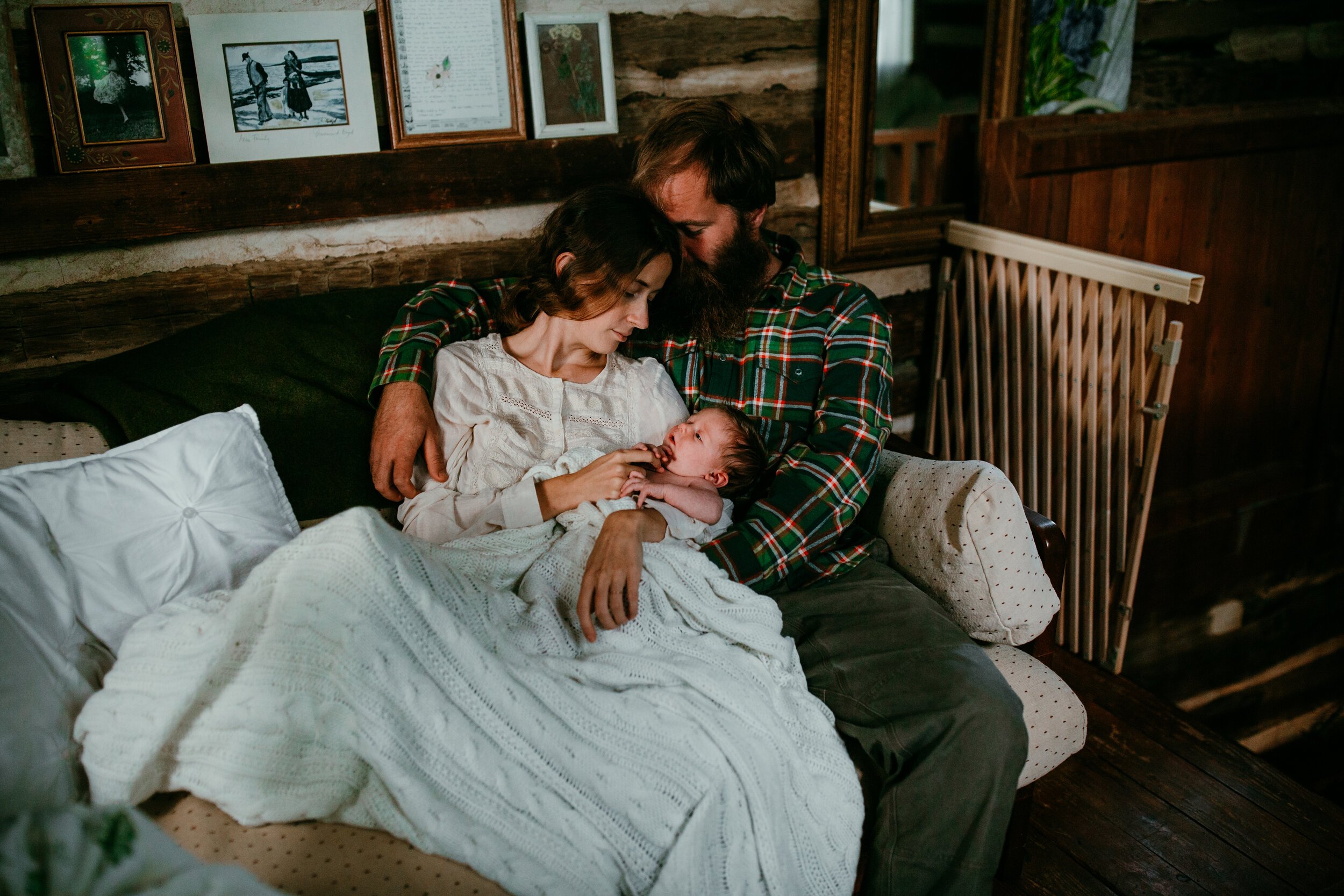 couple sits under blanket in their log cabin holding brand new baby 