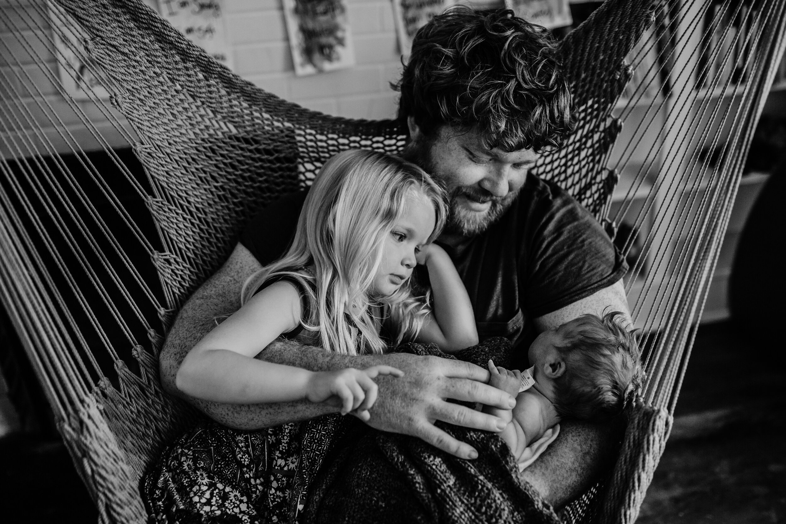 father sitting in hammock looks at newborn daughter while also holding toddler big sister 
