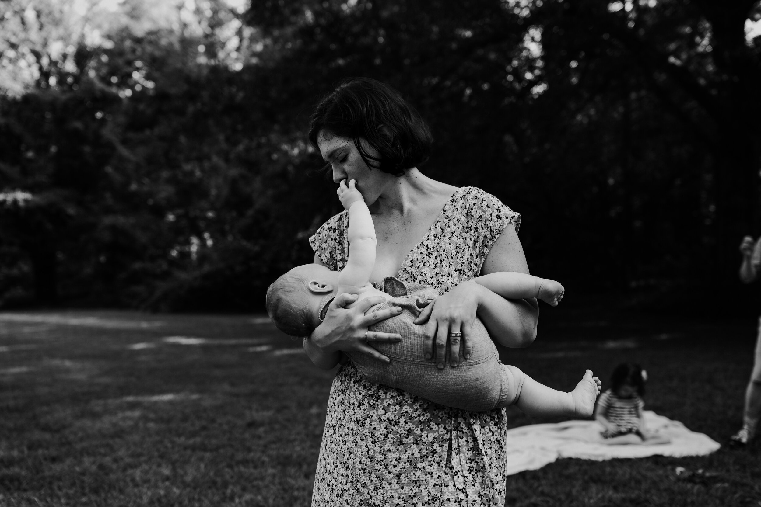 mother nurses infant son standing up in a field with family playing behind her in black and white 