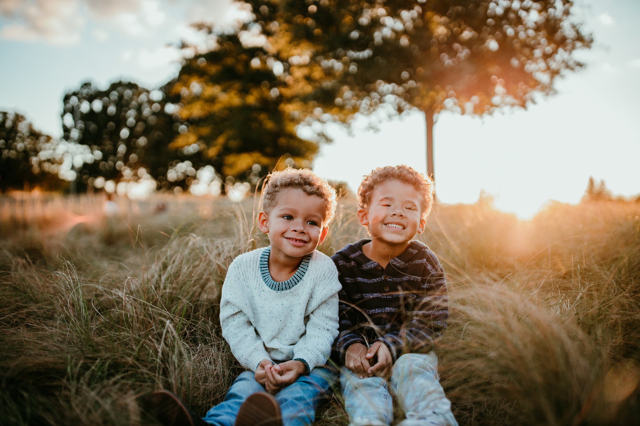 brothers sit together in field smiling at the camera with sunset in the background