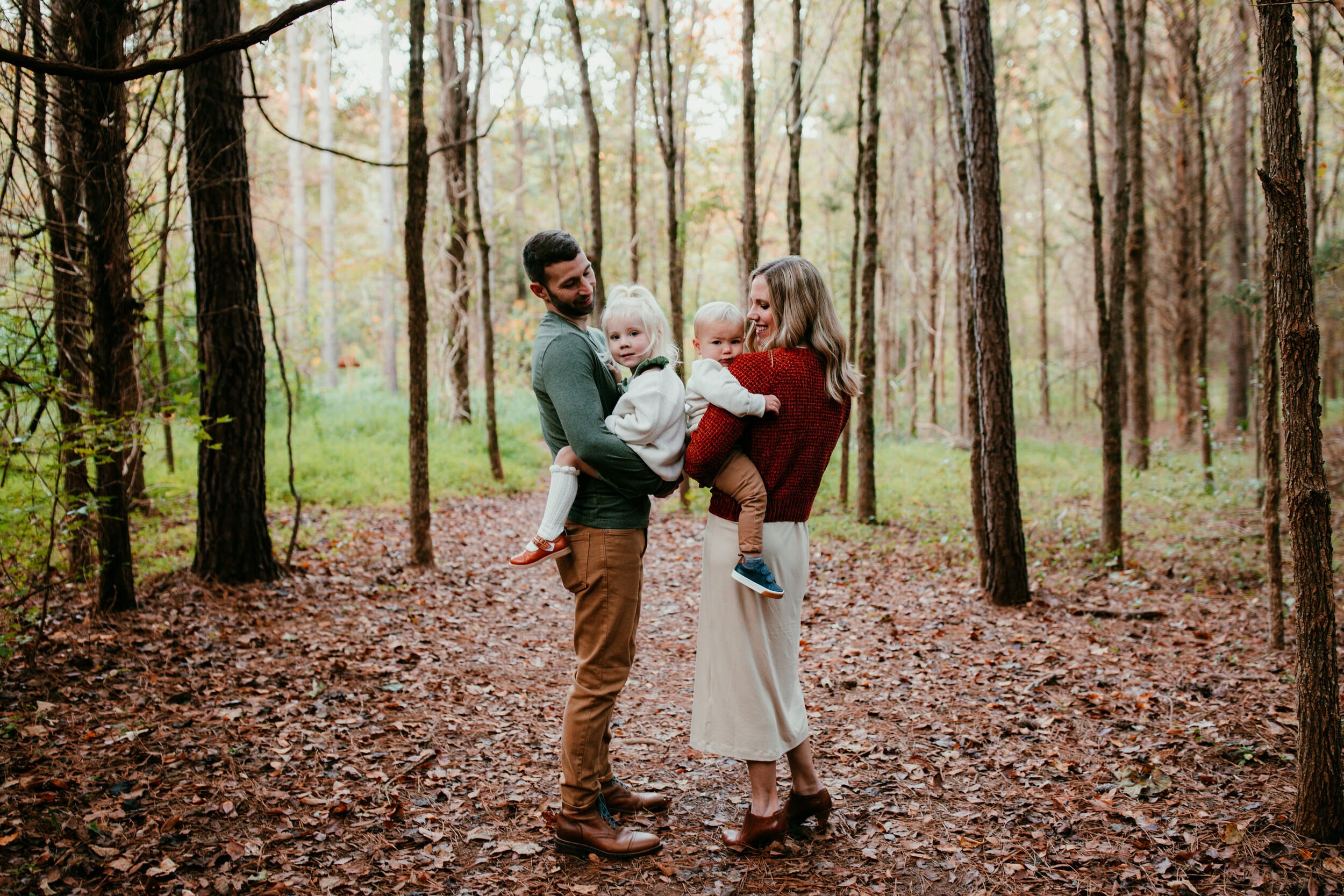 family of four smile together in the woods surrounded by Fall leaves