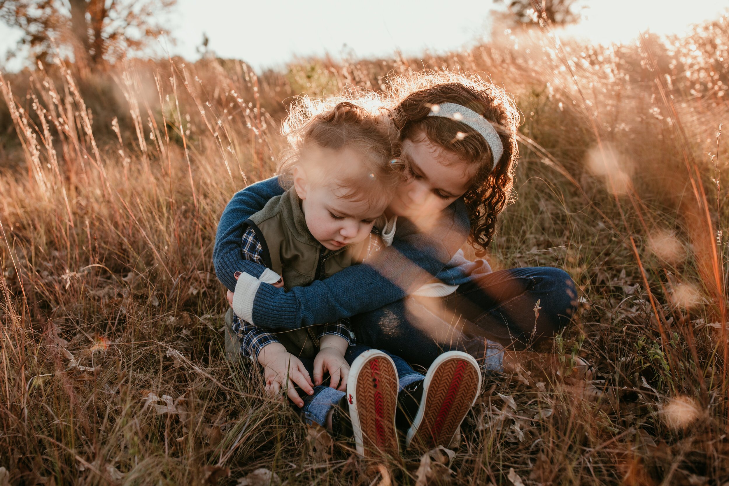 two siblings snuggle while sitting in a field at sunset amidst tall grass