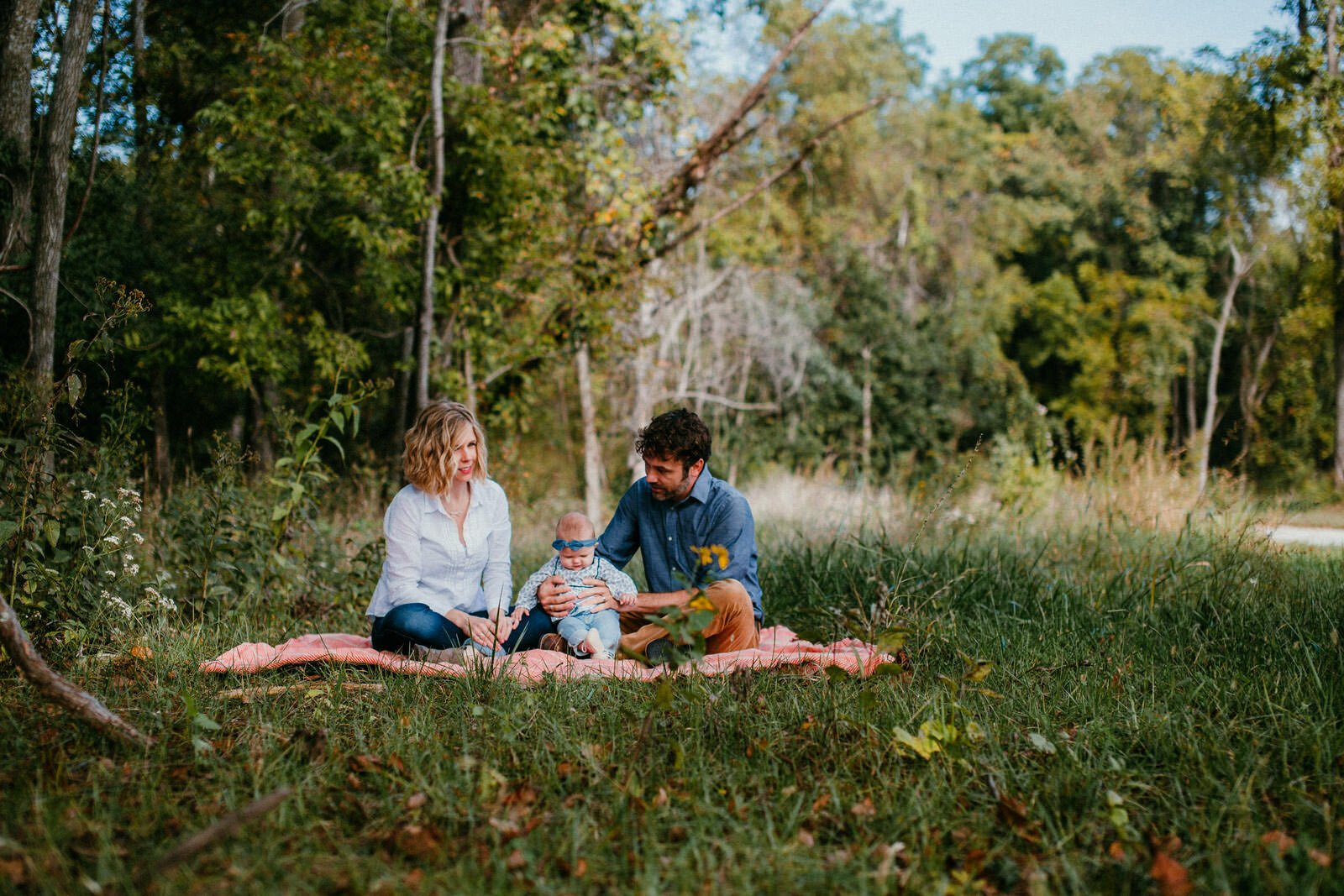 family of three sit together on pink picnic blanket in an overgrown green fields