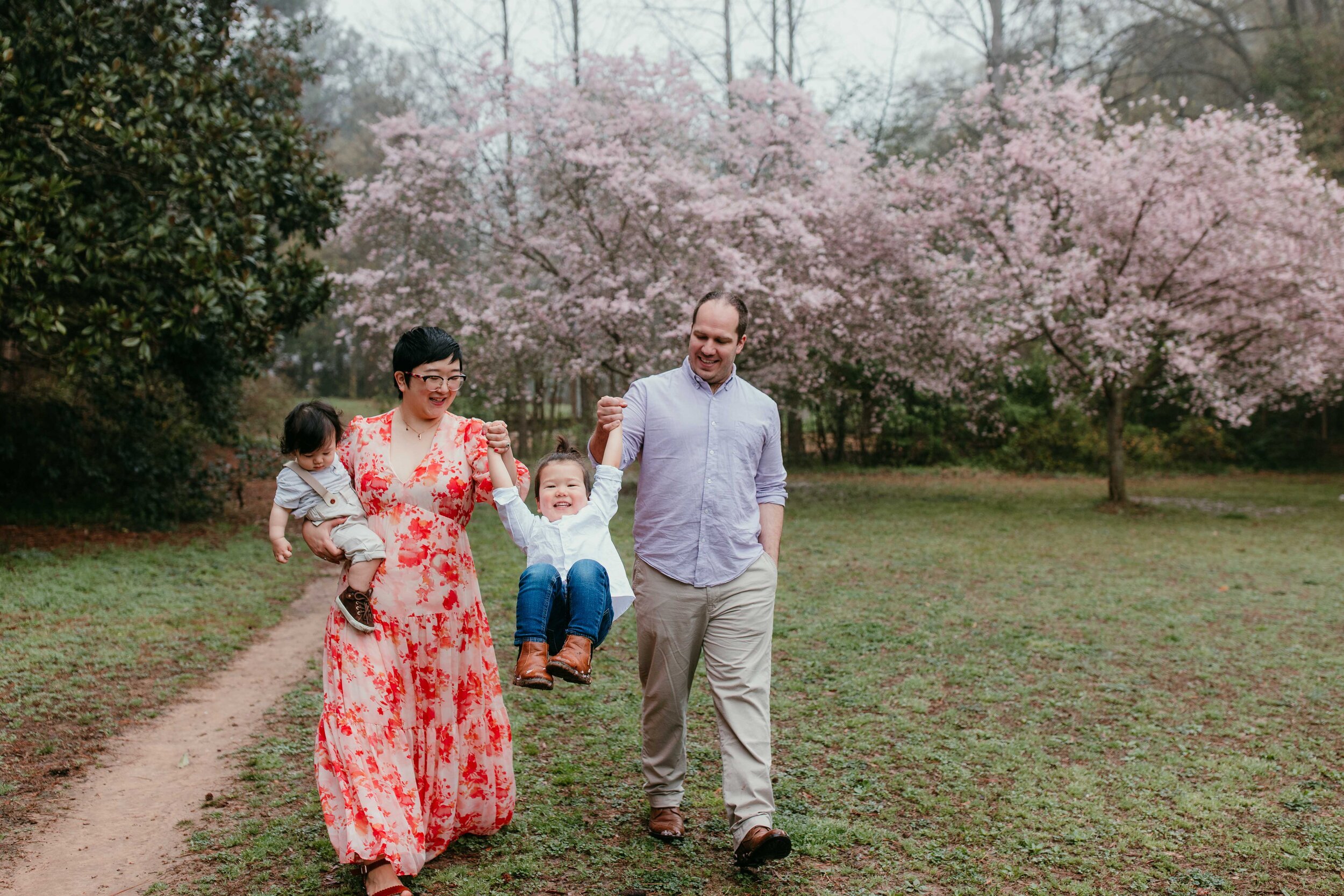 family of four walk together in grass field in front of pink flowering trees in Spring 