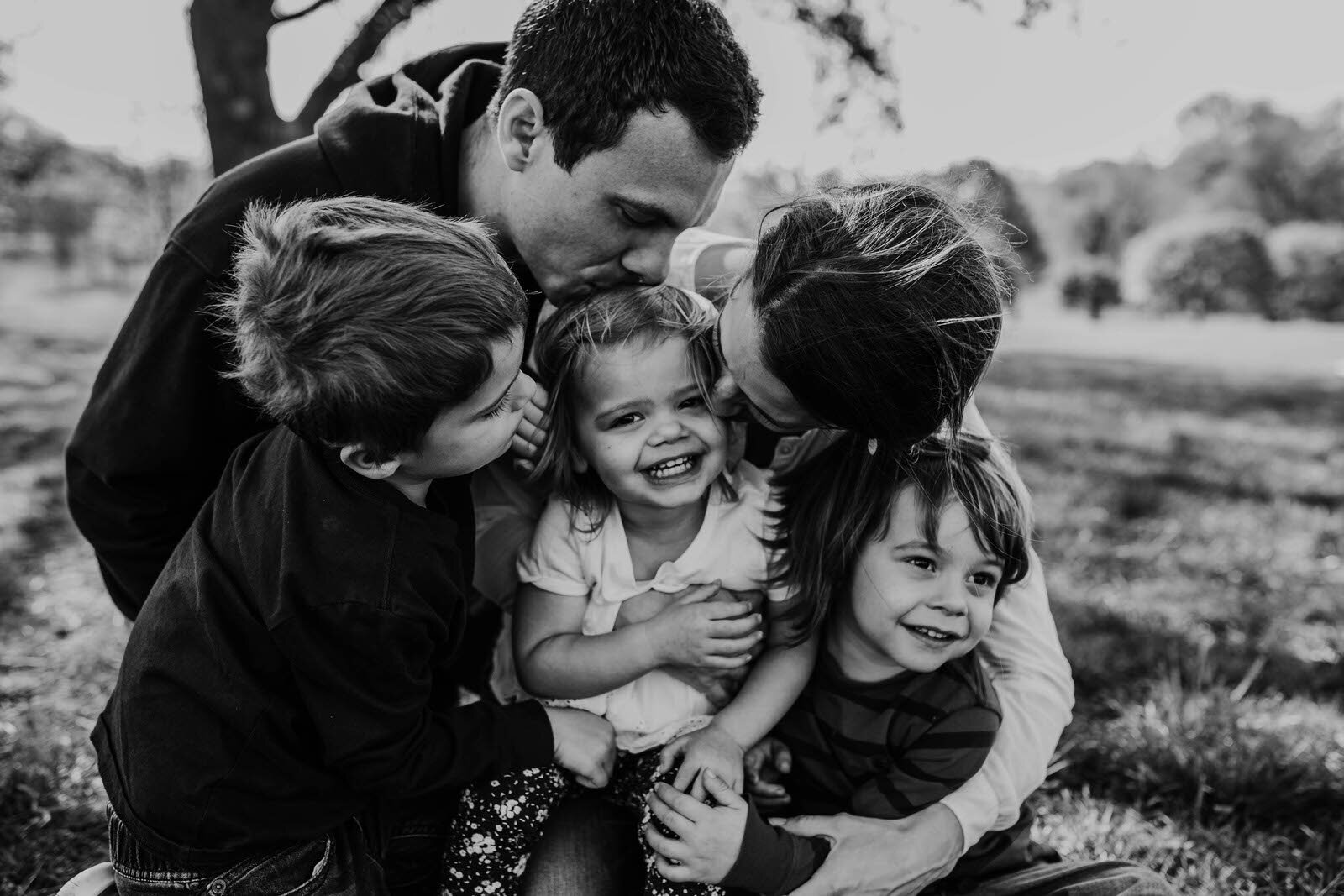 family of 5 snuggle together all leaning in to kiss youngest daughter in the middle of the hug black and white