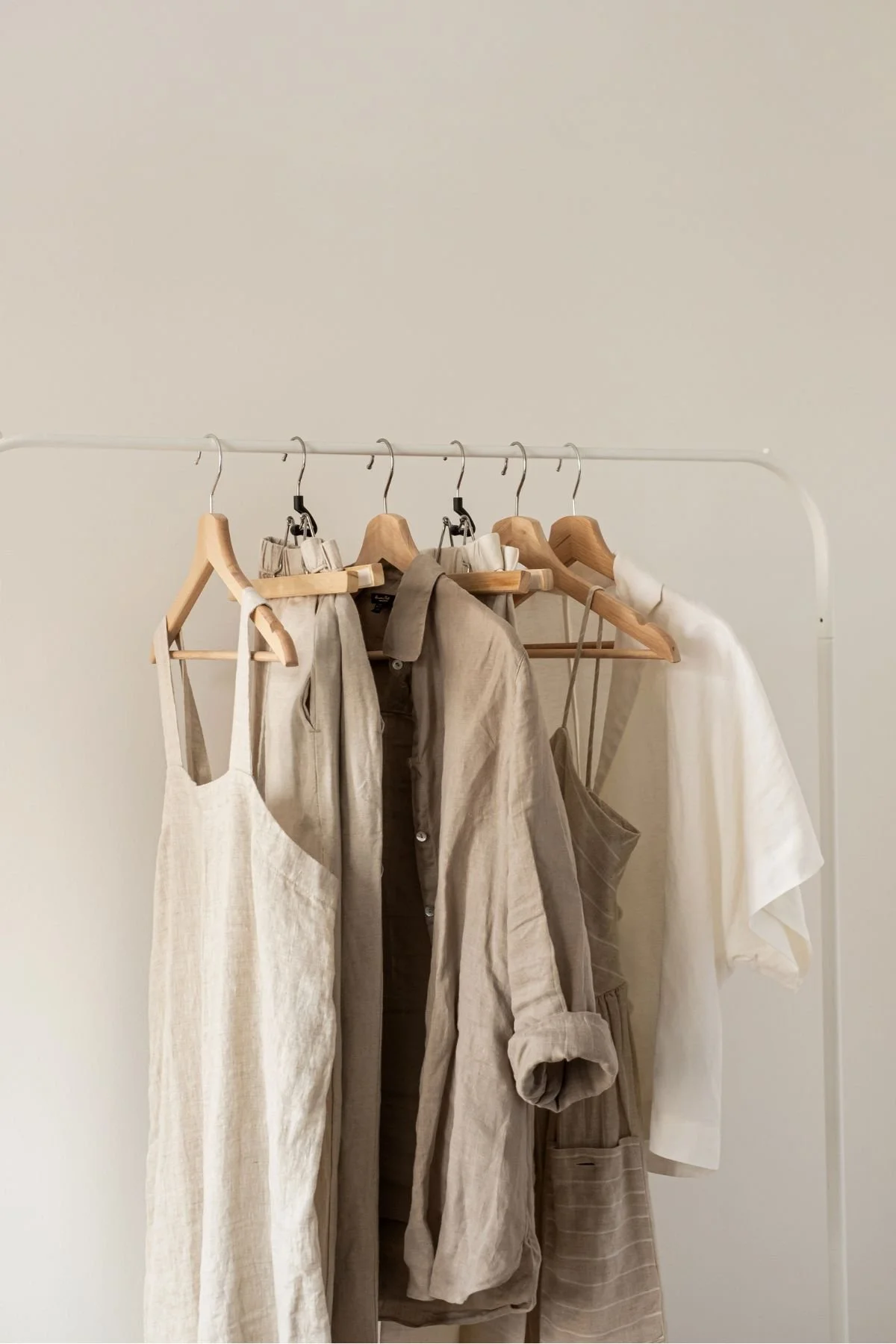 Clothing rack with beige and white casual linen garments including dresses, shirts, and a jacket on wooden and metal hangers against a plain light-colored wall.