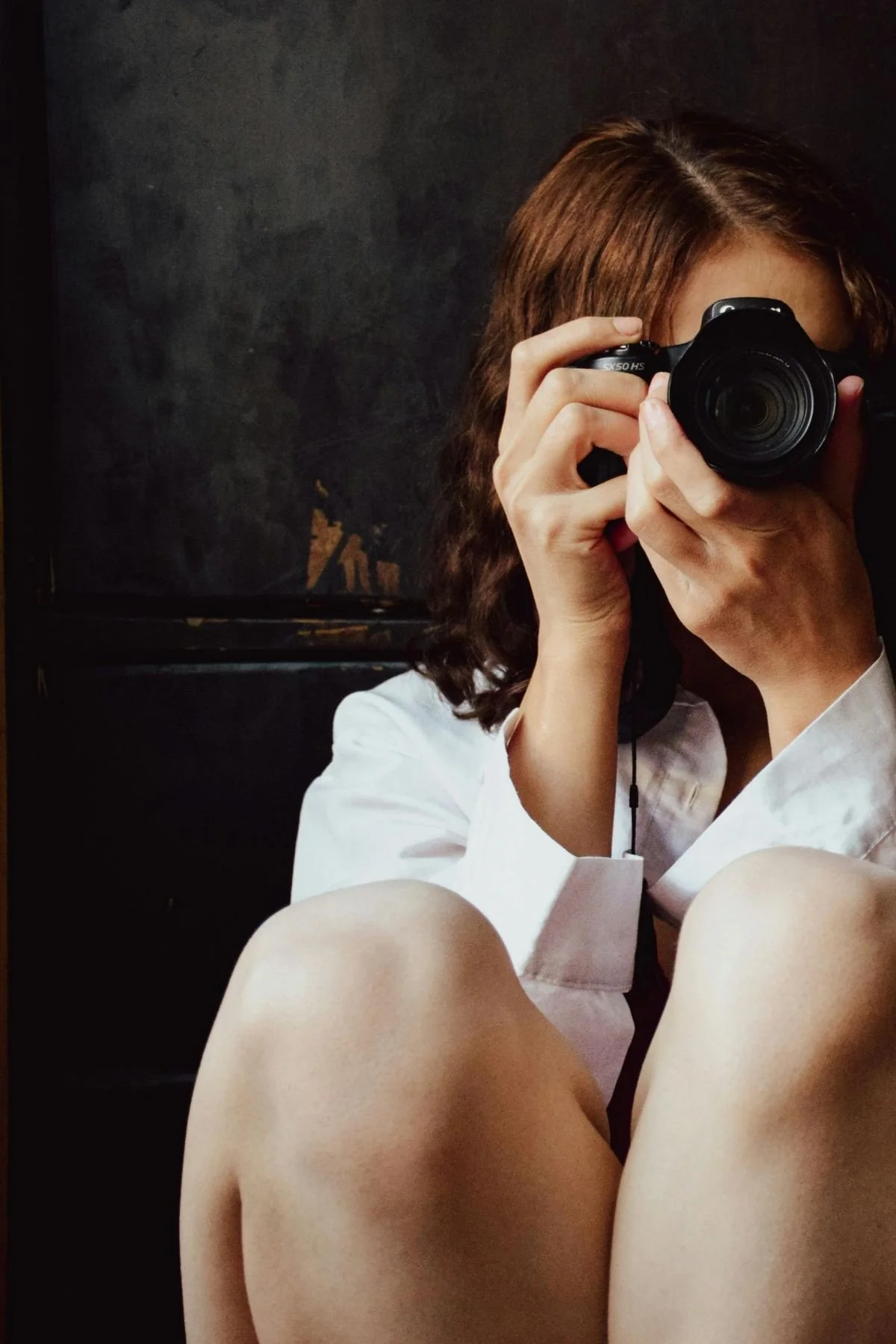 A woman with brown hair taking a photo with a digital camera, wearing a white shirt, sitting against a dark background.