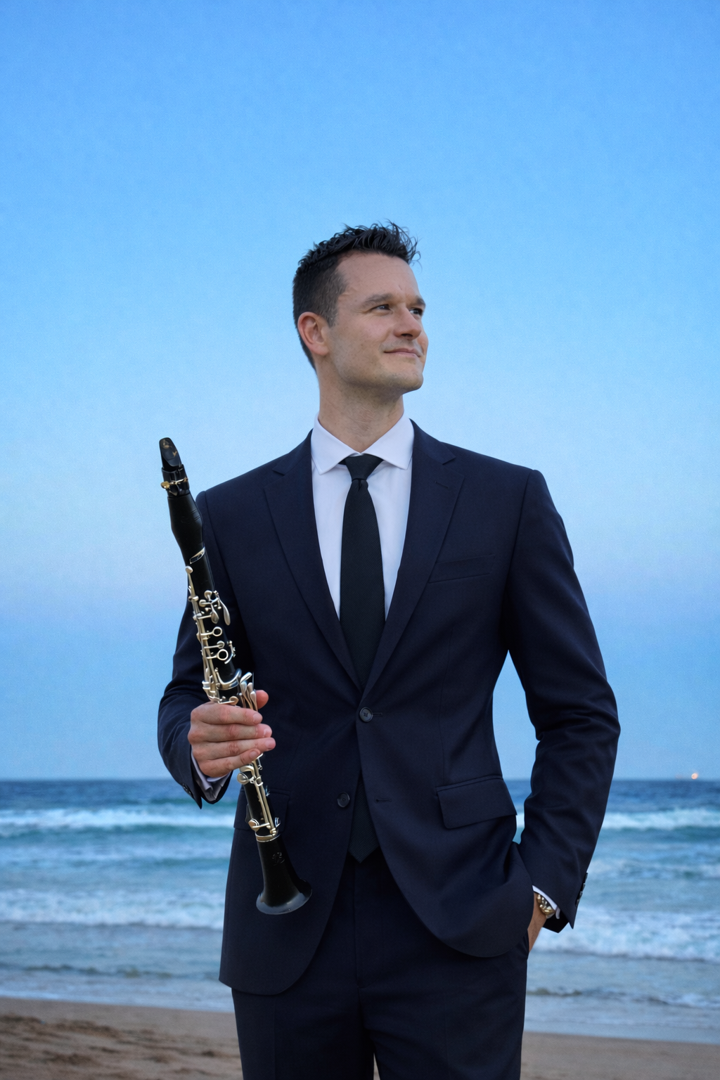 A man in a dark suit holding a clarinet standing on a beach with the ocean in the background.