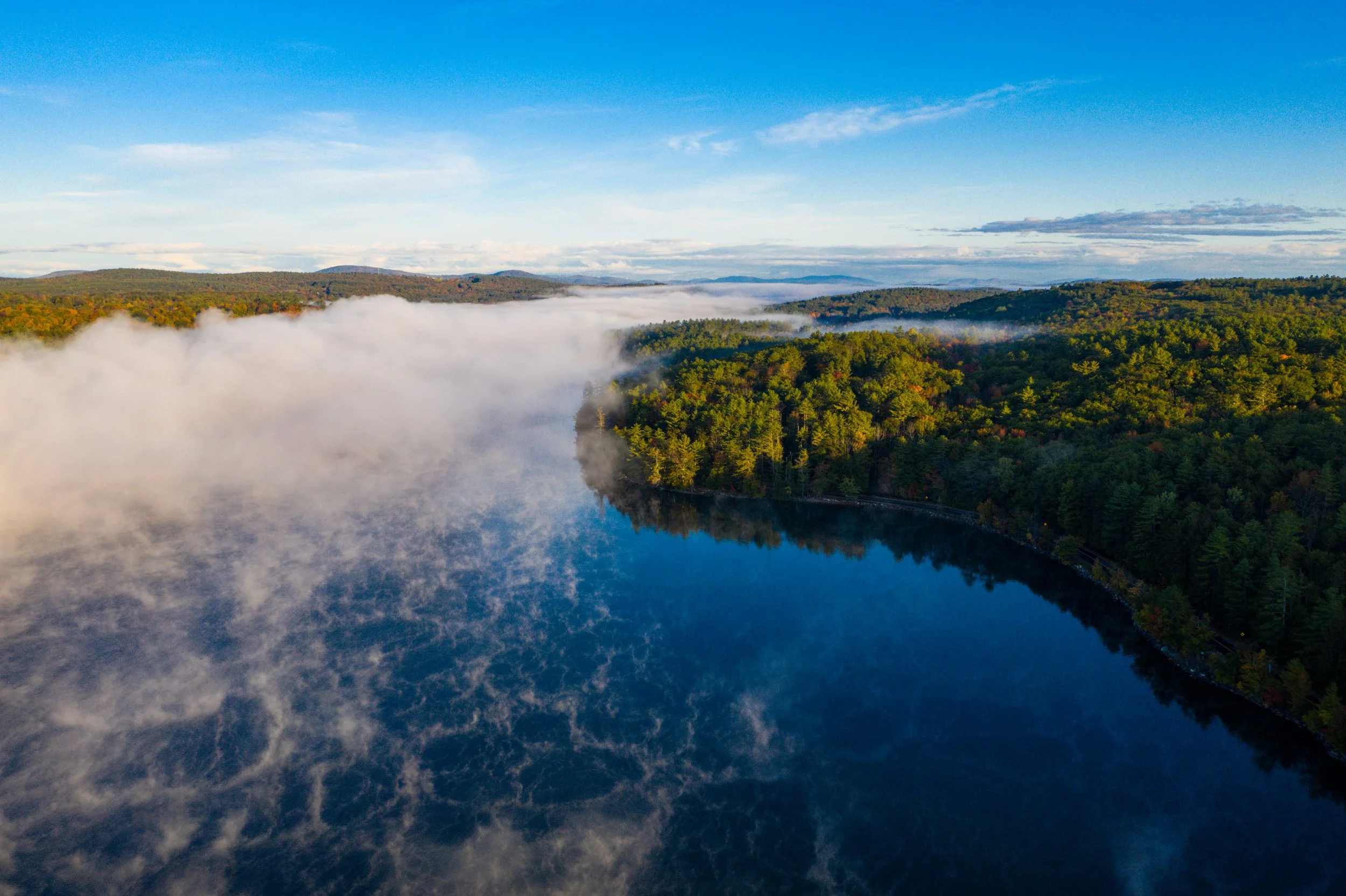 Aerial view of a Maine lake surrounded by dense forest with mist rising from the water and a clear blue sky.