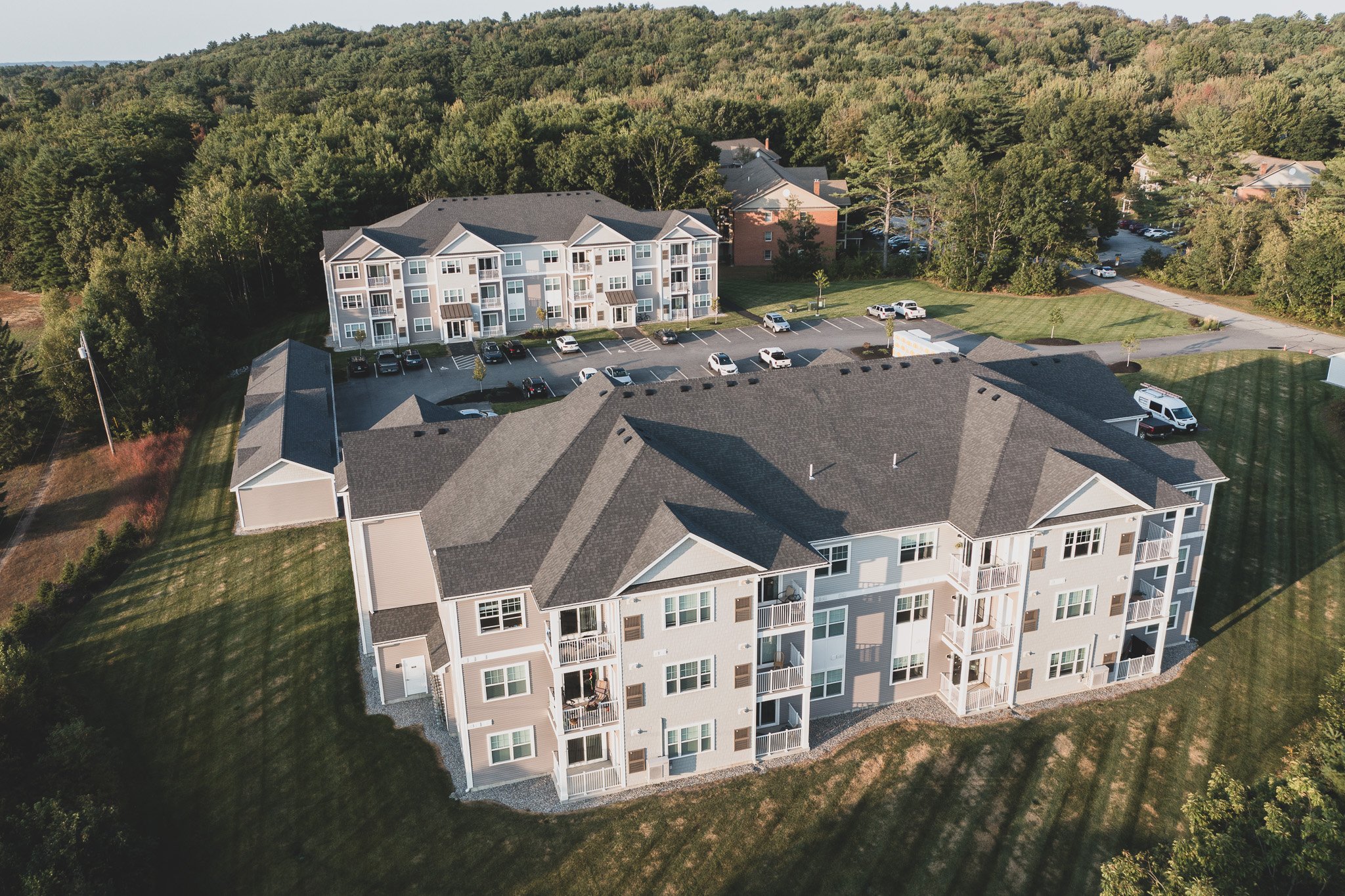 Aerial view of an apartment complex in Auburn, Maine surrounded by grass and trees, featuring two buildings with three stories, parking lots, and several parked vehicles.