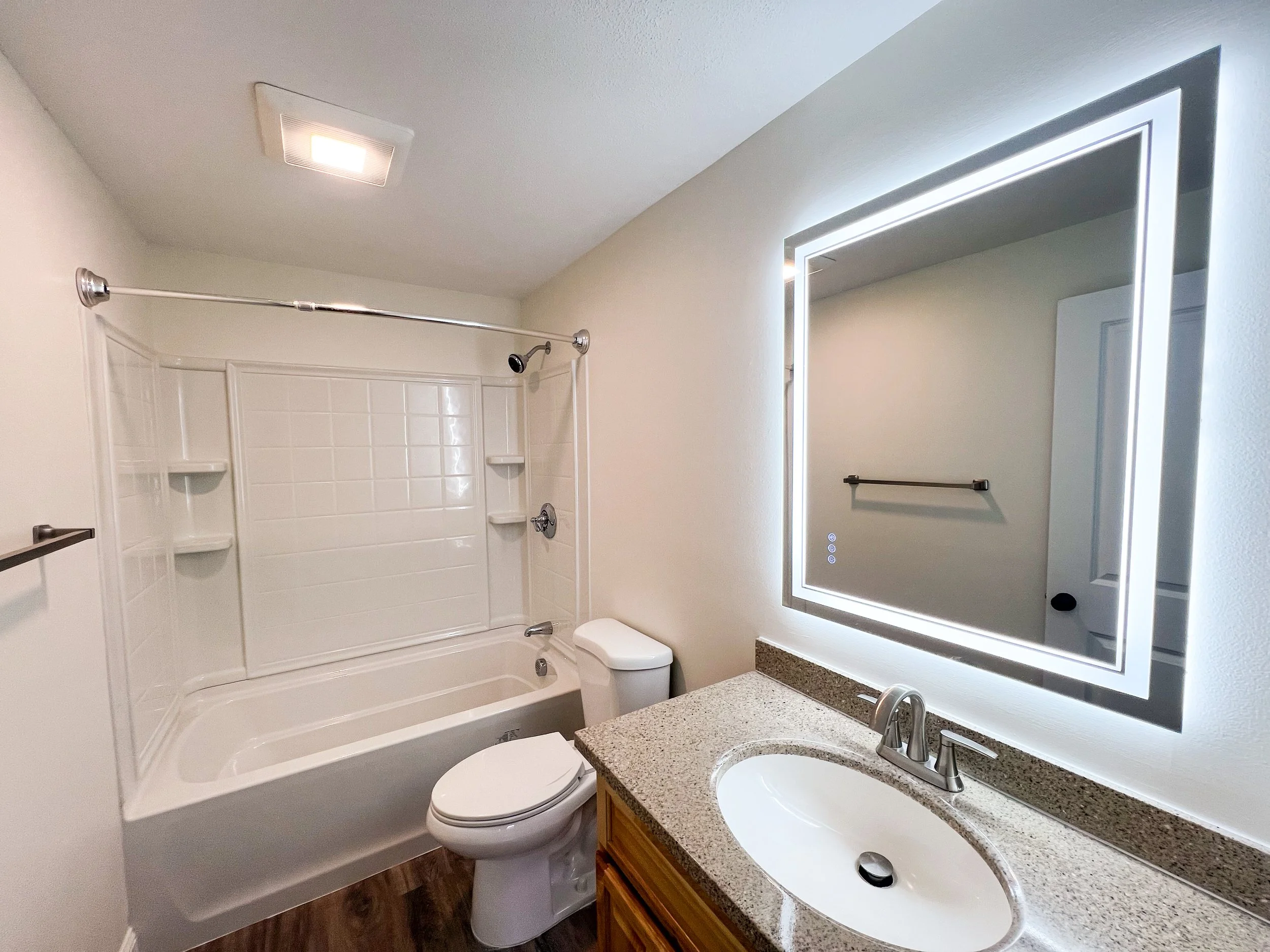 Modern bathroom with a bathtub and shower combo, a toilet, a sink with a granite countertop, and a backlit mirror.