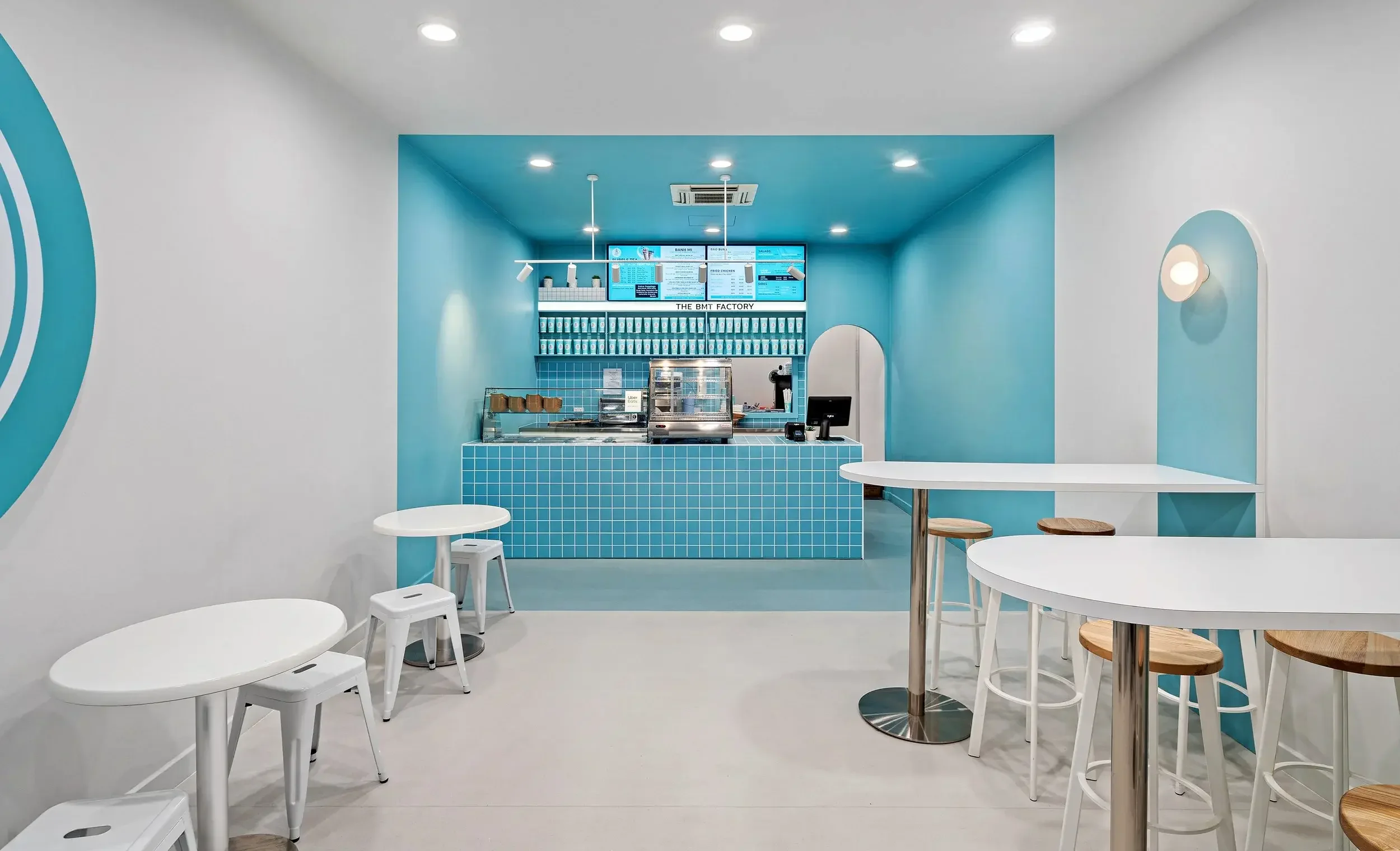 Modern cafe with white and blue decor, round tables with stools, and a counter with a digital menu display.