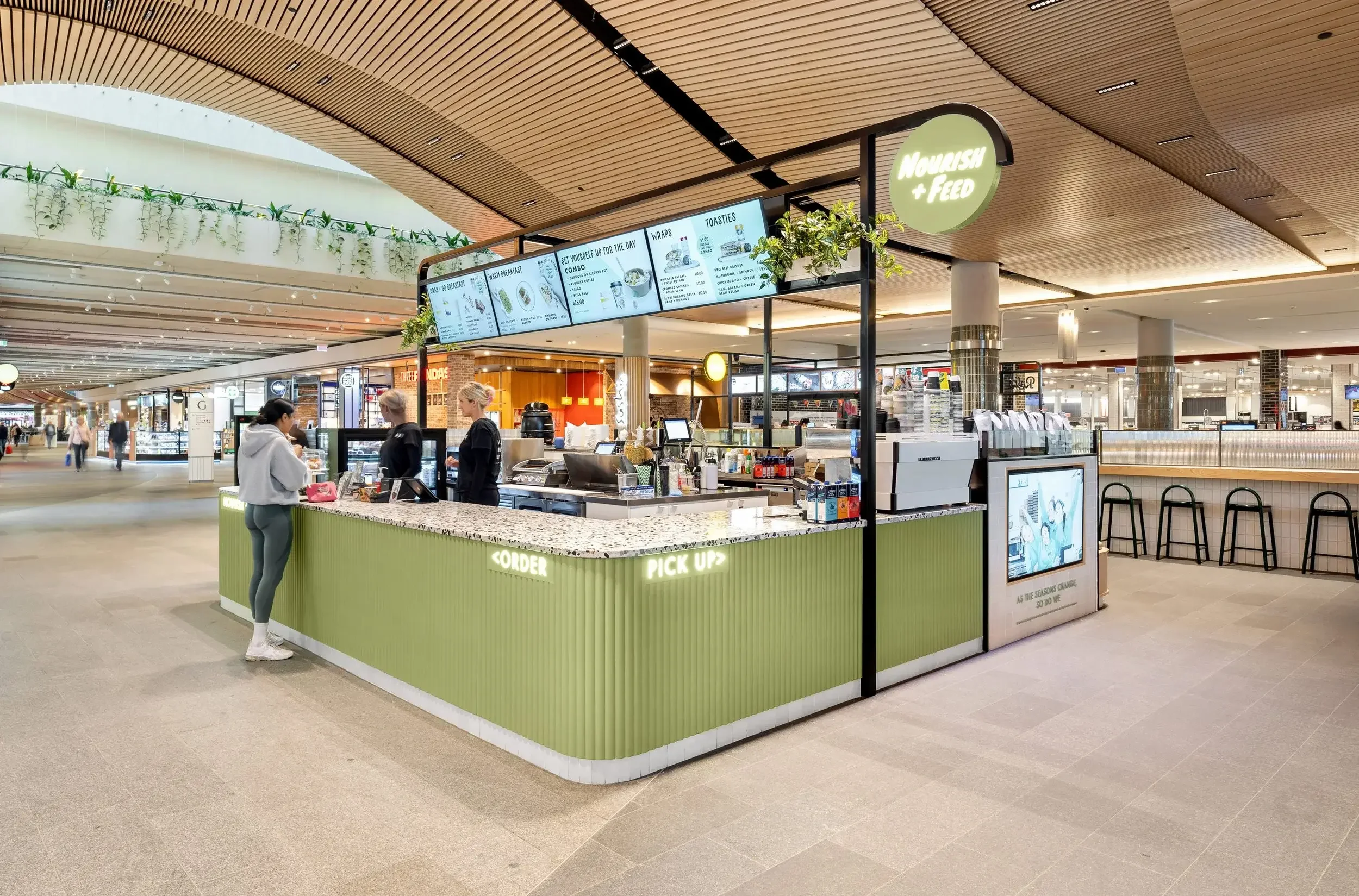 A food kiosk in an airport with a green exterior, digital menu boards, and staff preparing orders. Customers are ordering and picking up food.