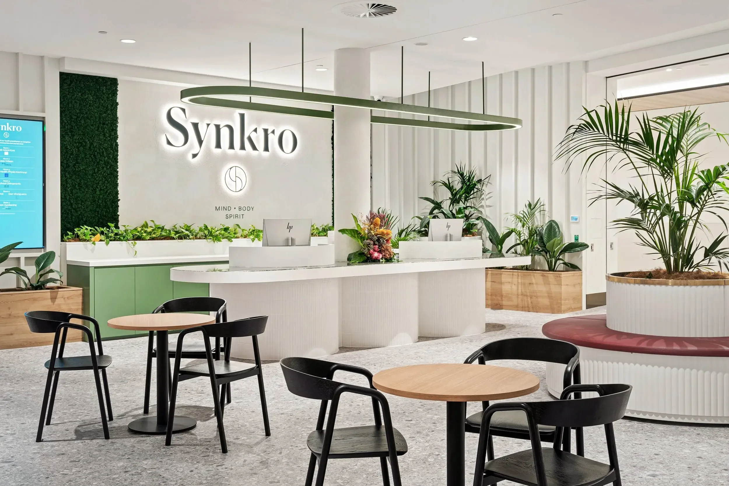 Modern office reception area with the sign 'Synkro' illuminated on the wall, surrounded by green plants, a white curved reception desk with two computer monitors, a bouquet of flowers, and small round tables with black chairs.