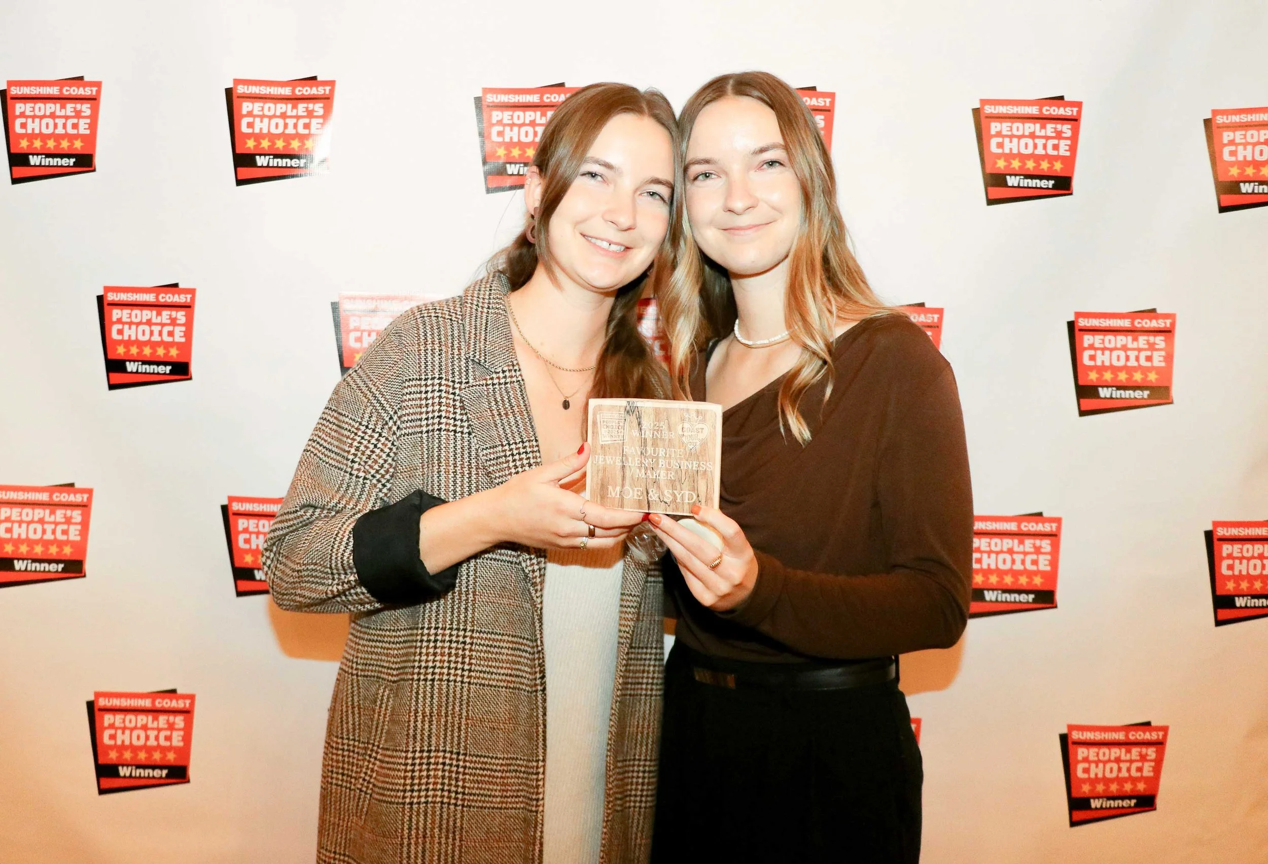 Two women smiling and holding a wooden plaque at a Sunshine Coast People's Choice awards event, with a backdrop featuring multiple red and black award logos.