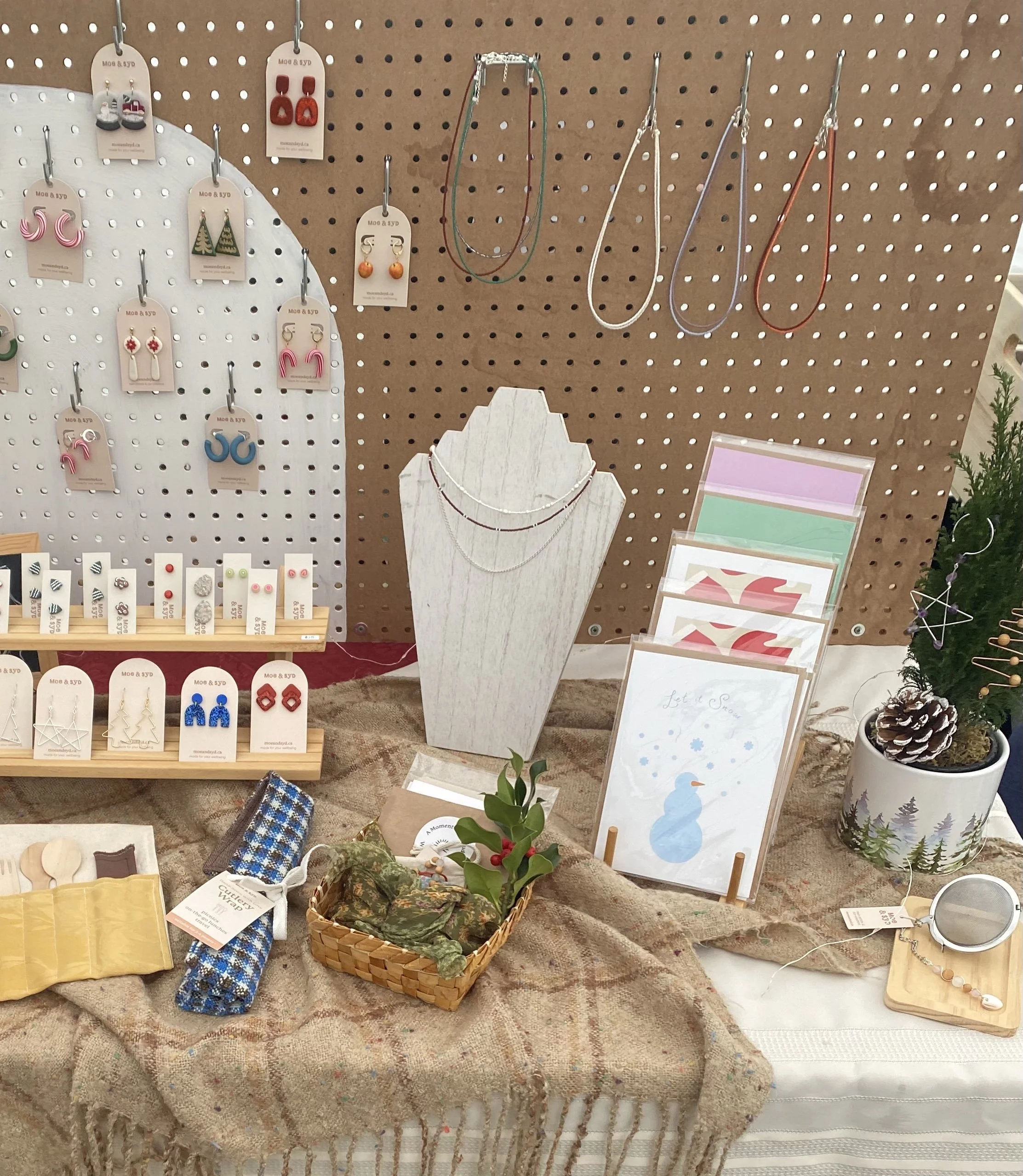 Display of jewelry and accessories including necklaces, earrings, and pins, arranged on a pegboard and table with decorative items like a small tree and pine cone, in a craft or boutique shop.