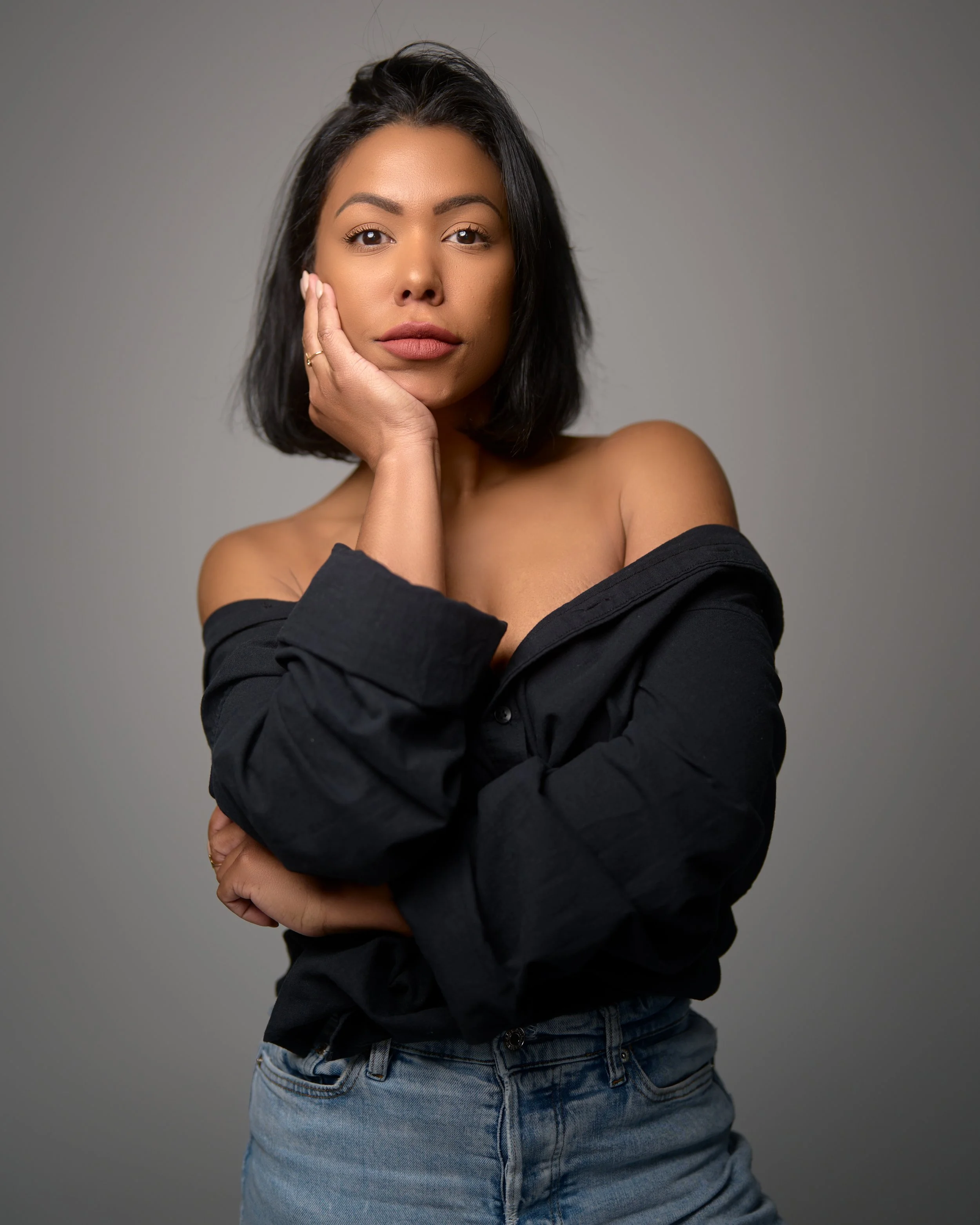 A young woman with black hair sitting on a green chair against a gray background, wearing a black blazer and black top with white cuffs, resting her head on her left hand and looking at the camera.
