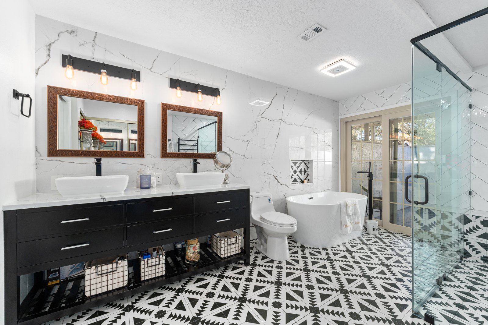 A modern bathroom vanity with a white cabinet, marble countertop, dual sinks, black faucets, and two large mirrors. Wall-mounted light fixtures with four bulbs each are above the mirrors. The wall is painted light gray, and a glass shower enclosure is visible in the reflection of the left mirror.