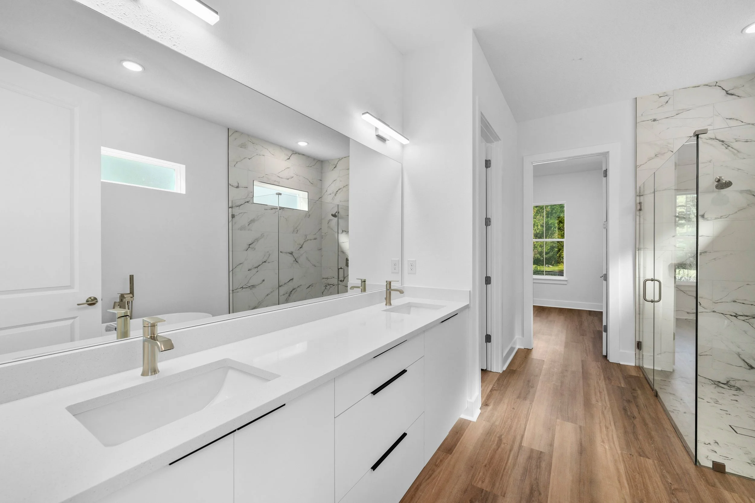 Modern bathroom with double white vanity, large mirror, gold faucets, wood flooring, and glass shower with marble tiles.