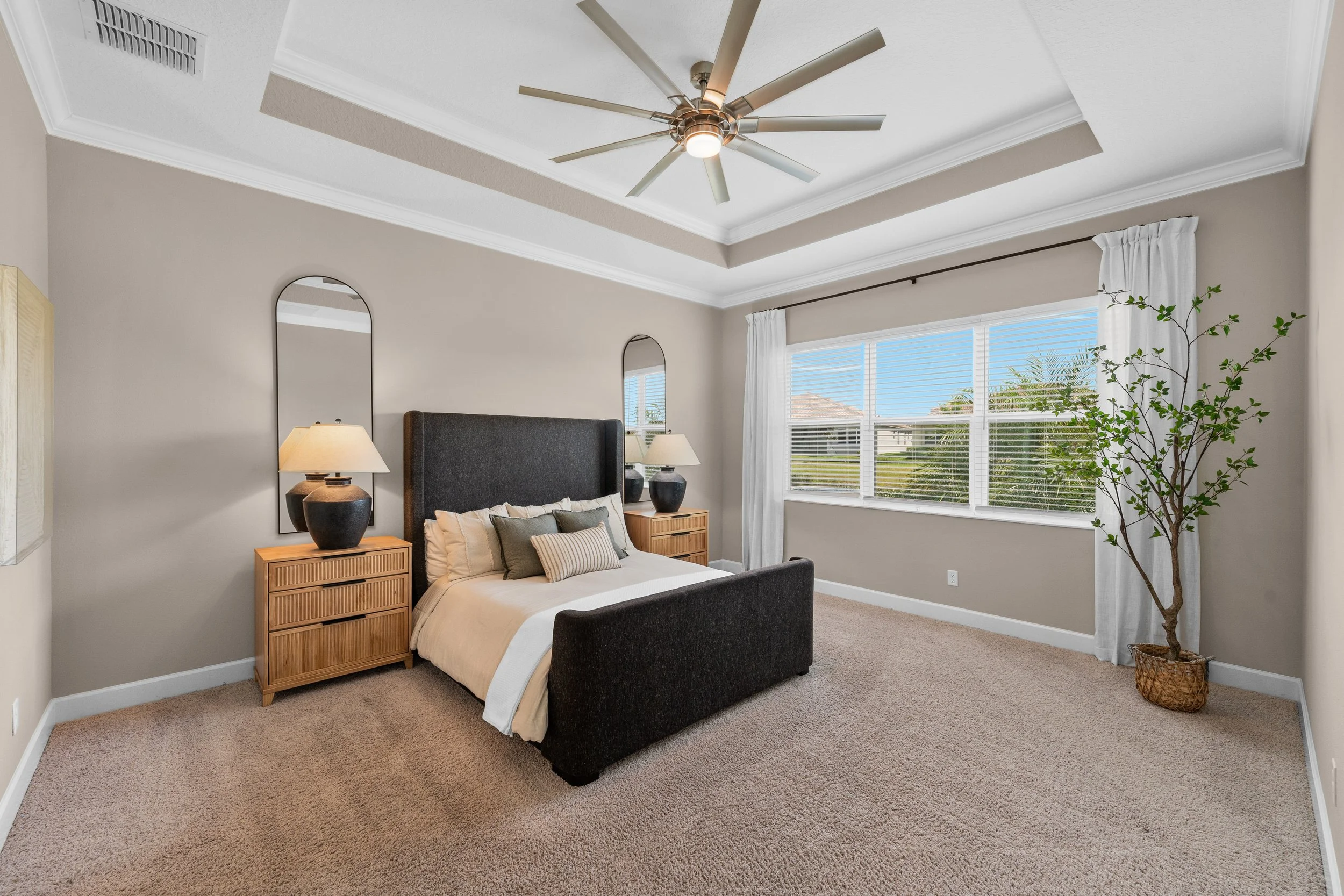A bedroom with a gray bed, two wooden nightstands with black lamps, and a large window with white curtains offering a view of a sunny yard with trees.