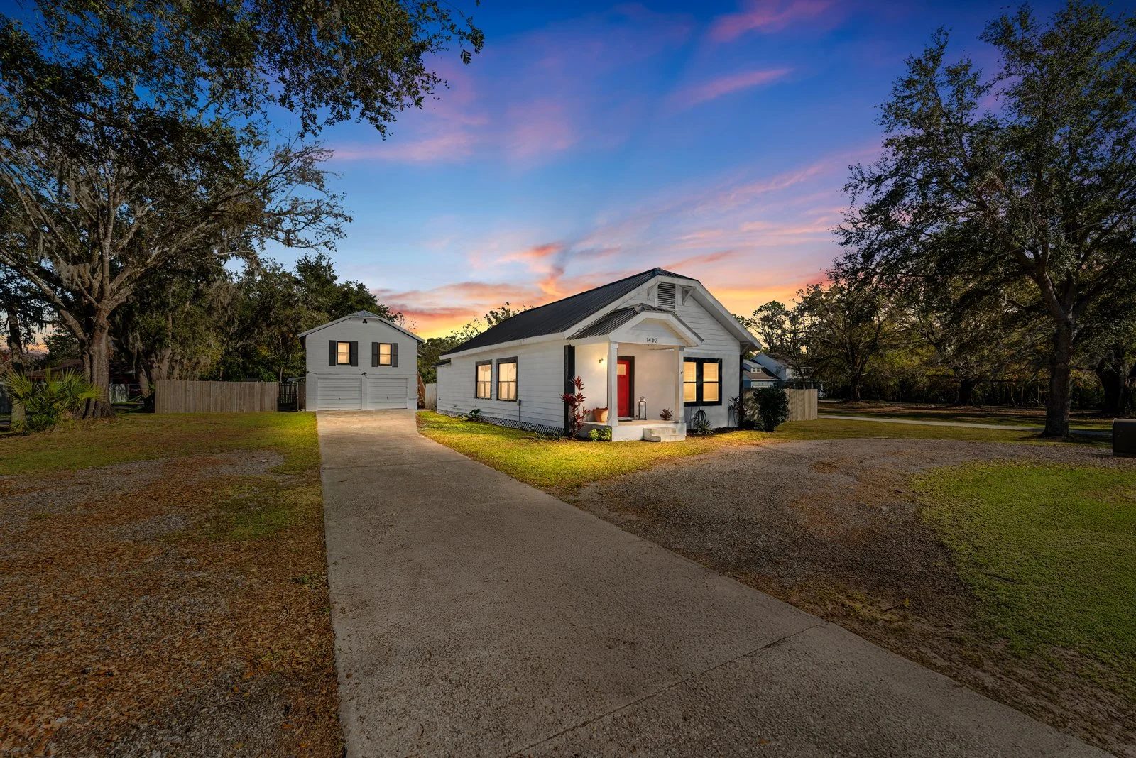 A charming white house with black window frames and a red door, located on a spacious lot with a driveway, surrounded by large trees, during sunset.