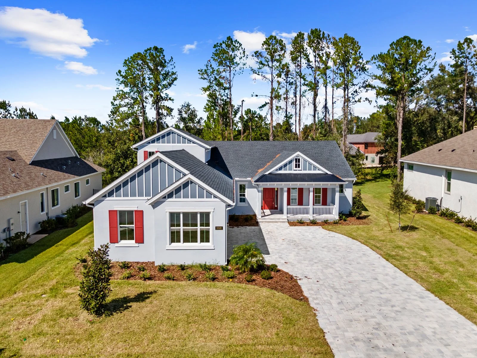 A modern two-story house with white walls, red shutters, and a gray roof, featuring a front porch with white railing and a paved driveway. The property is surrounded by green grass and neighboring houses, with tall trees in the background under a blue sky.