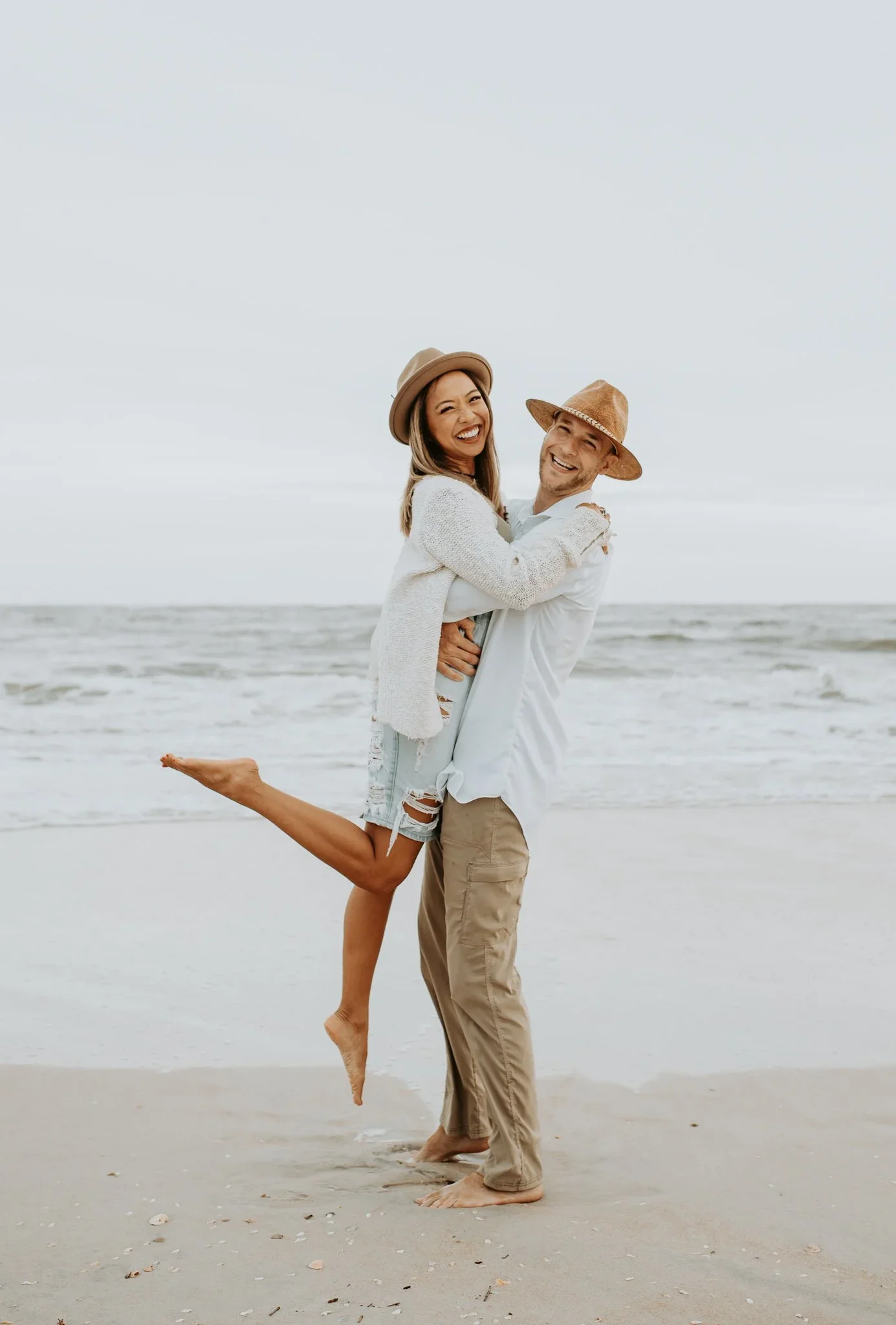A man lifting a woman on his shoulders on the beach, both smiling and wearing hats, with overcast skies in the background.