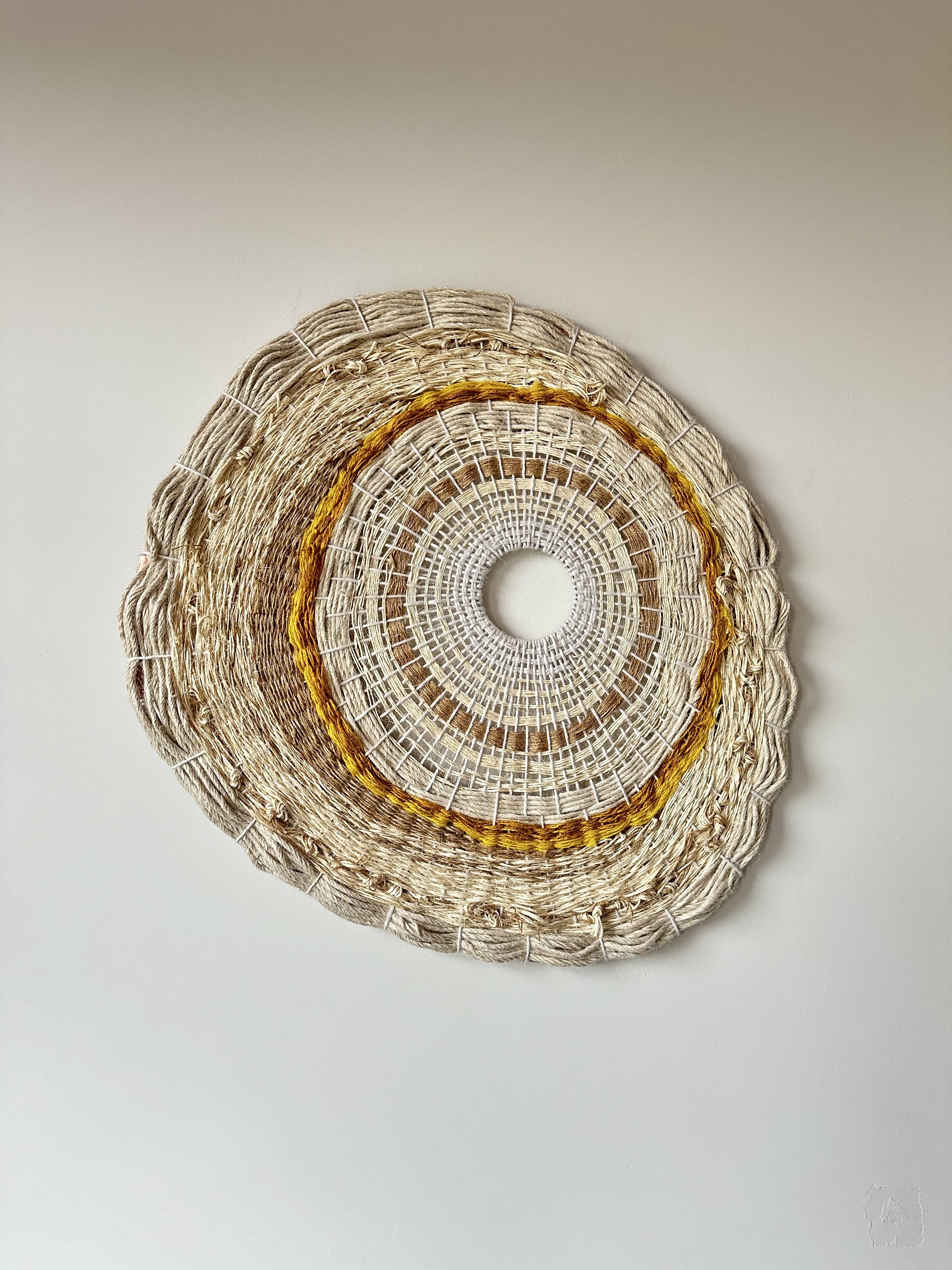 A circular woven wall hanging made of natural fibers in beige, brown, and gold tones, displayed on a plain white wall.