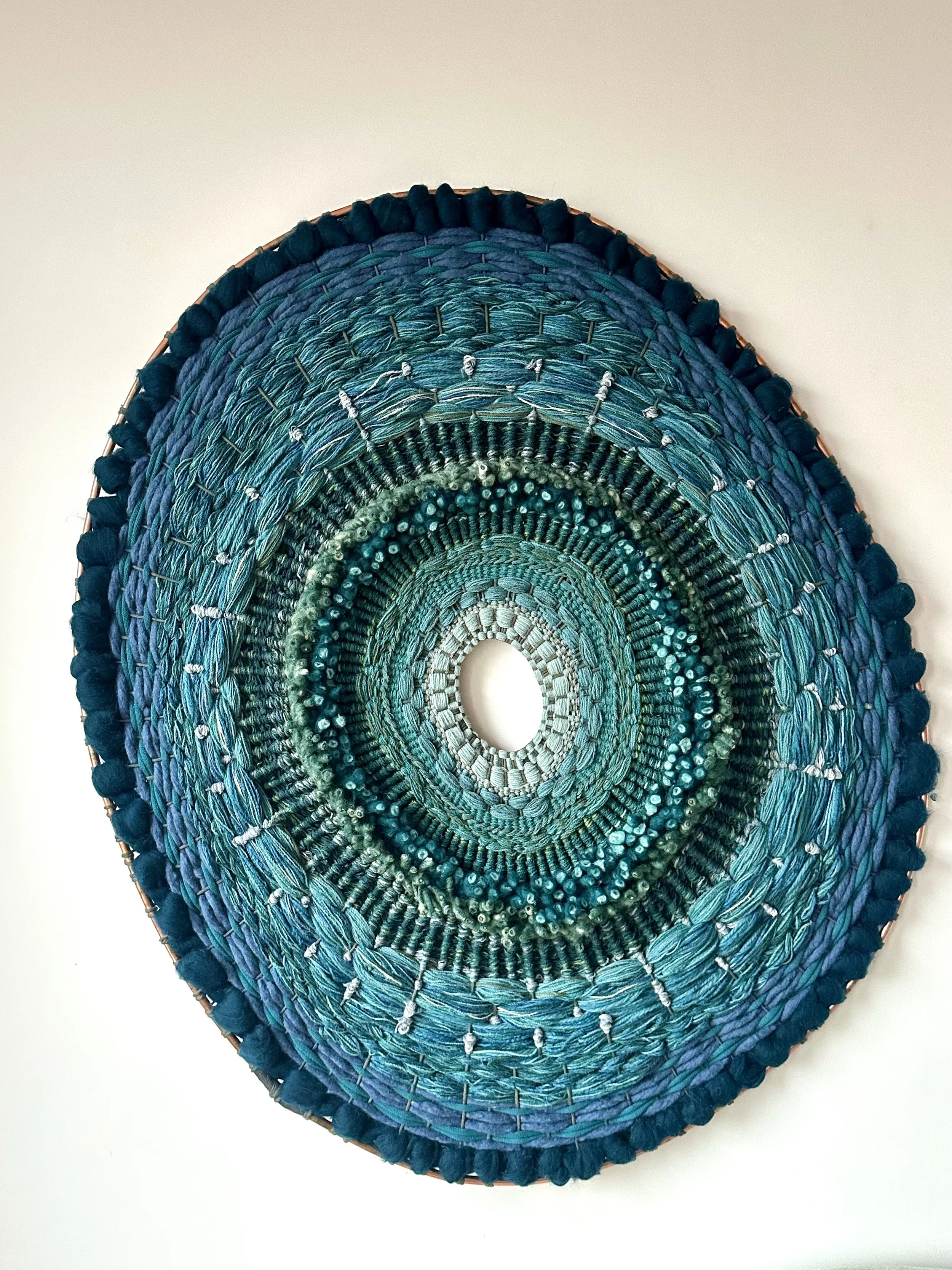 Hand-woven circular textile art piece in shades of blue and green with a central opening, displaying intricate patterns and textures on a wall.