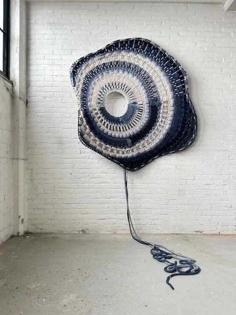 A large, round woven textile artwork with blue and white concentric patterns hangs on a white brick wall. A long blue cord extends onto the floor from the center of the piece.