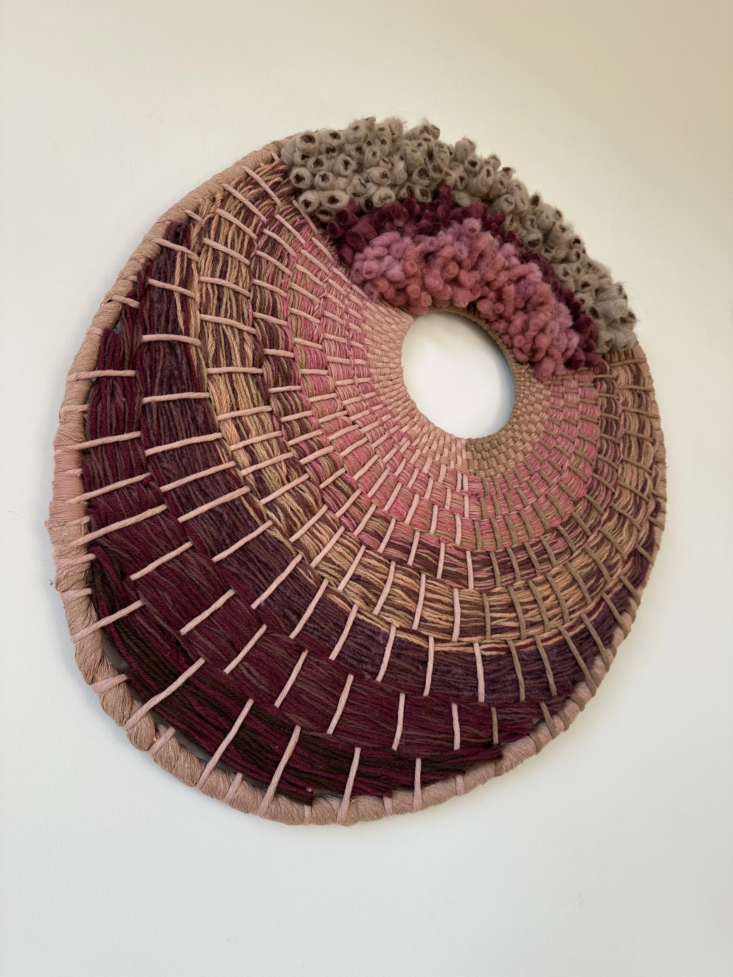 Circular woven wall art with varying shades of pink, purple, and beige yarn, featuring textured and layered fibers.
