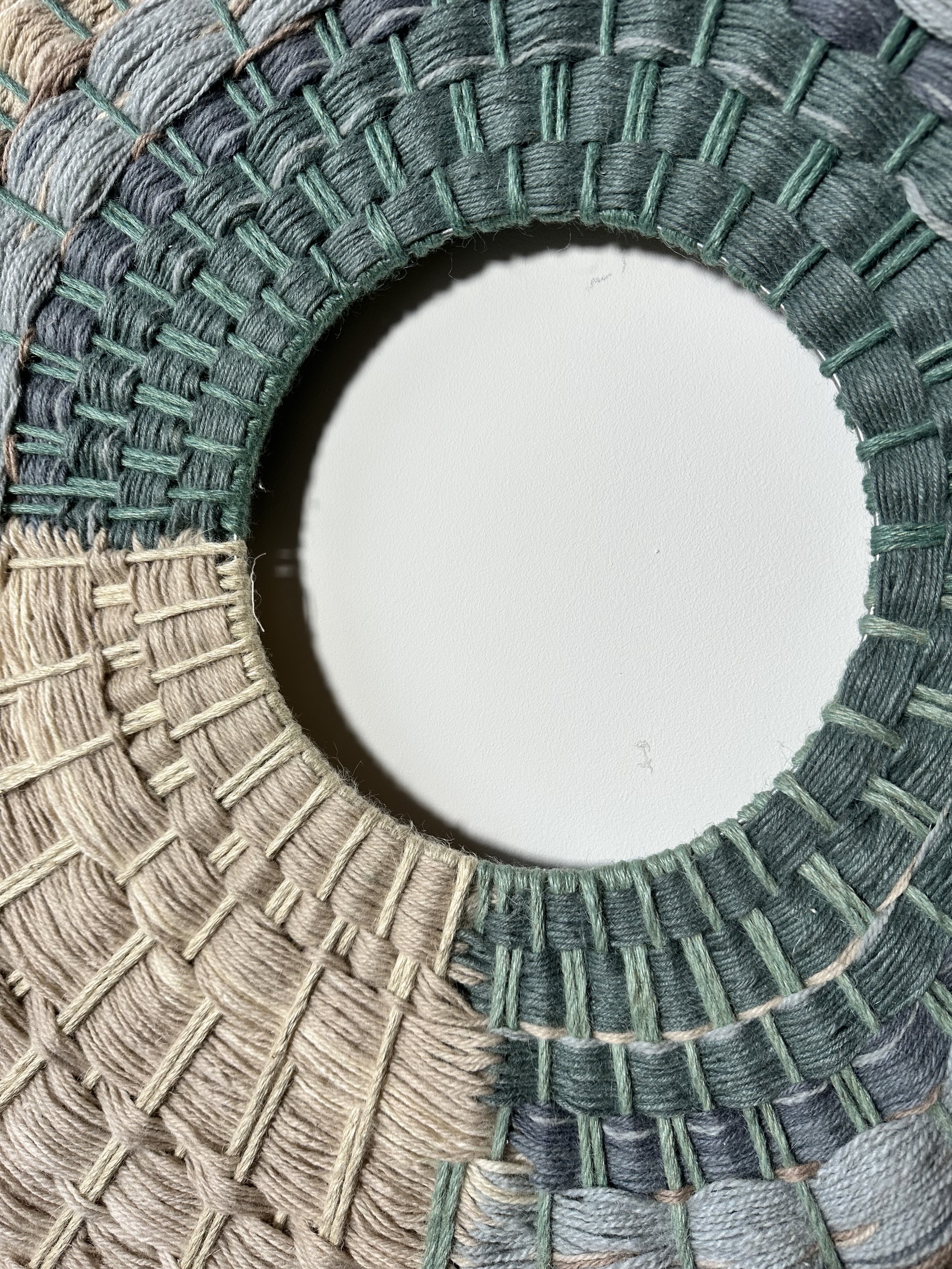 Close-up of a circular woven pattern in shades of green, gray, and beige, resembling a fiber art or macrame design.