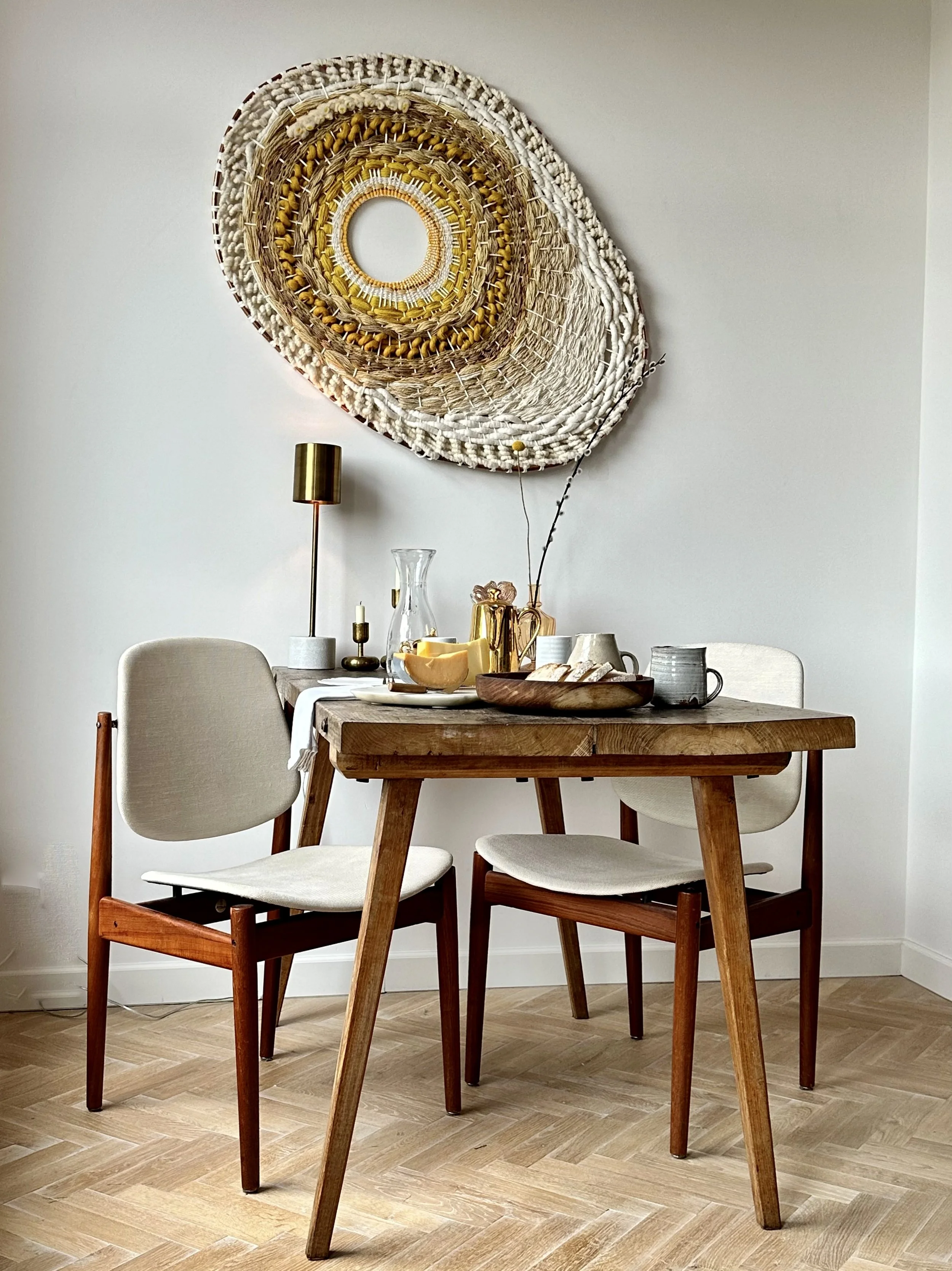 A dining area with a wooden table and two chairs featuring light-colored cushions. On the table are a water carafe, cups, a bowl, and some decorative items. Above the table hangs a large woven wall art piece with concentric circles in earthy tones. T