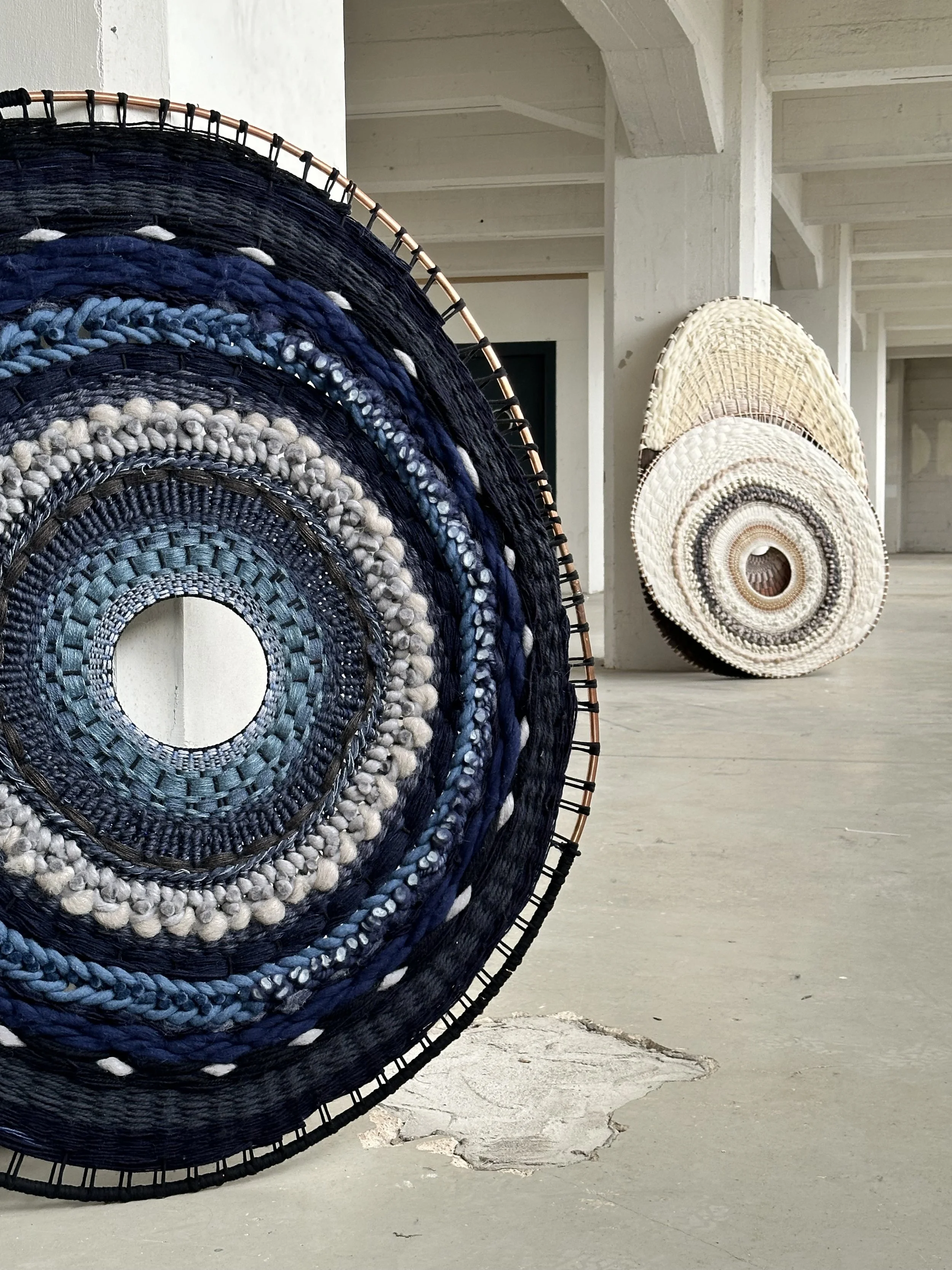 Large circular woven tapestries with intricate patterns, featuring shades of blue and neutral colors, displayed in an industrial-style space.