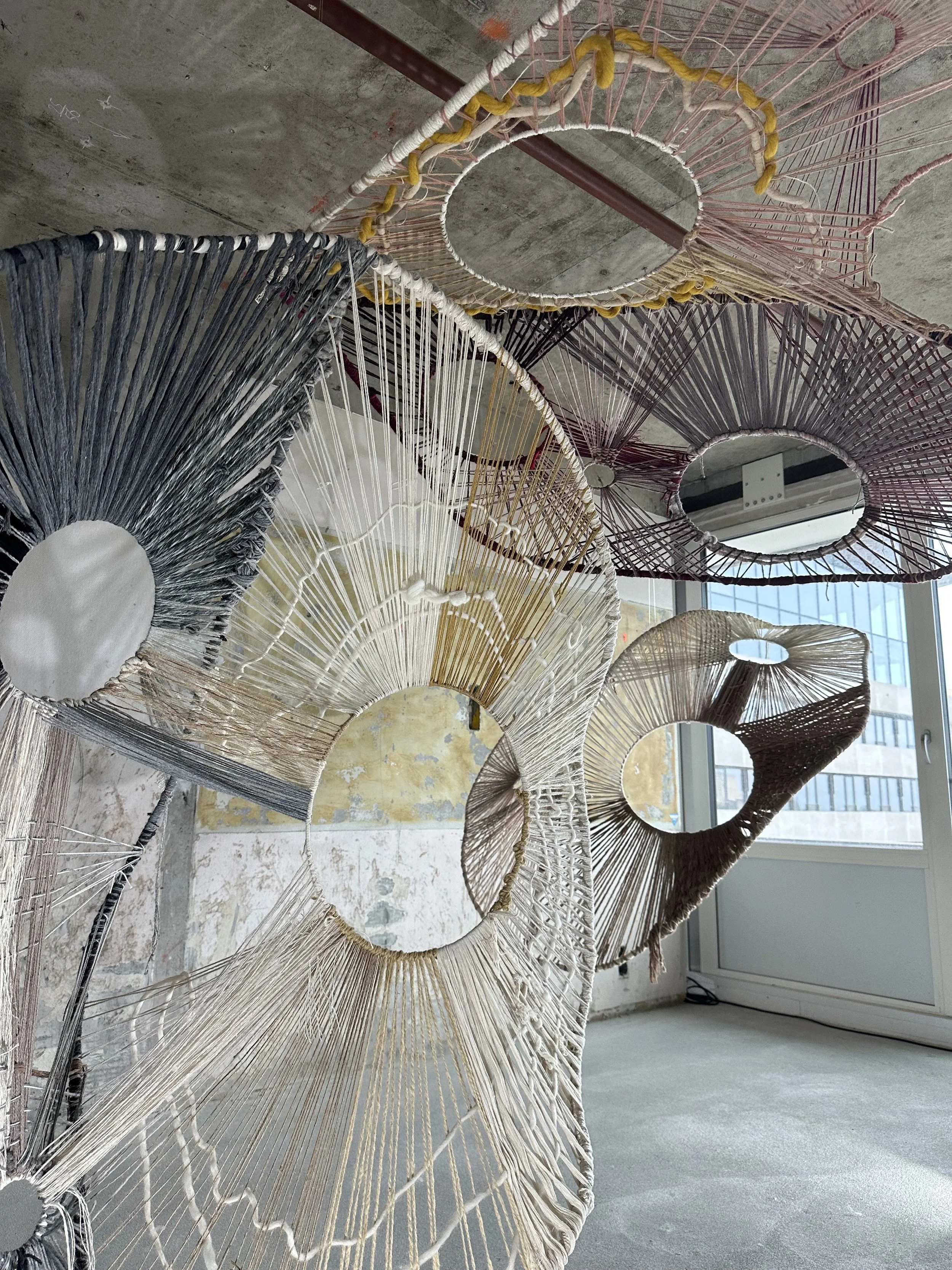 Art installation featuring circular frames with woven yarn patterns in a room with concrete ceiling and large windows. The yarn is arranged in various neutral tones, creating an intricate design with open circles and lines radiating outward.