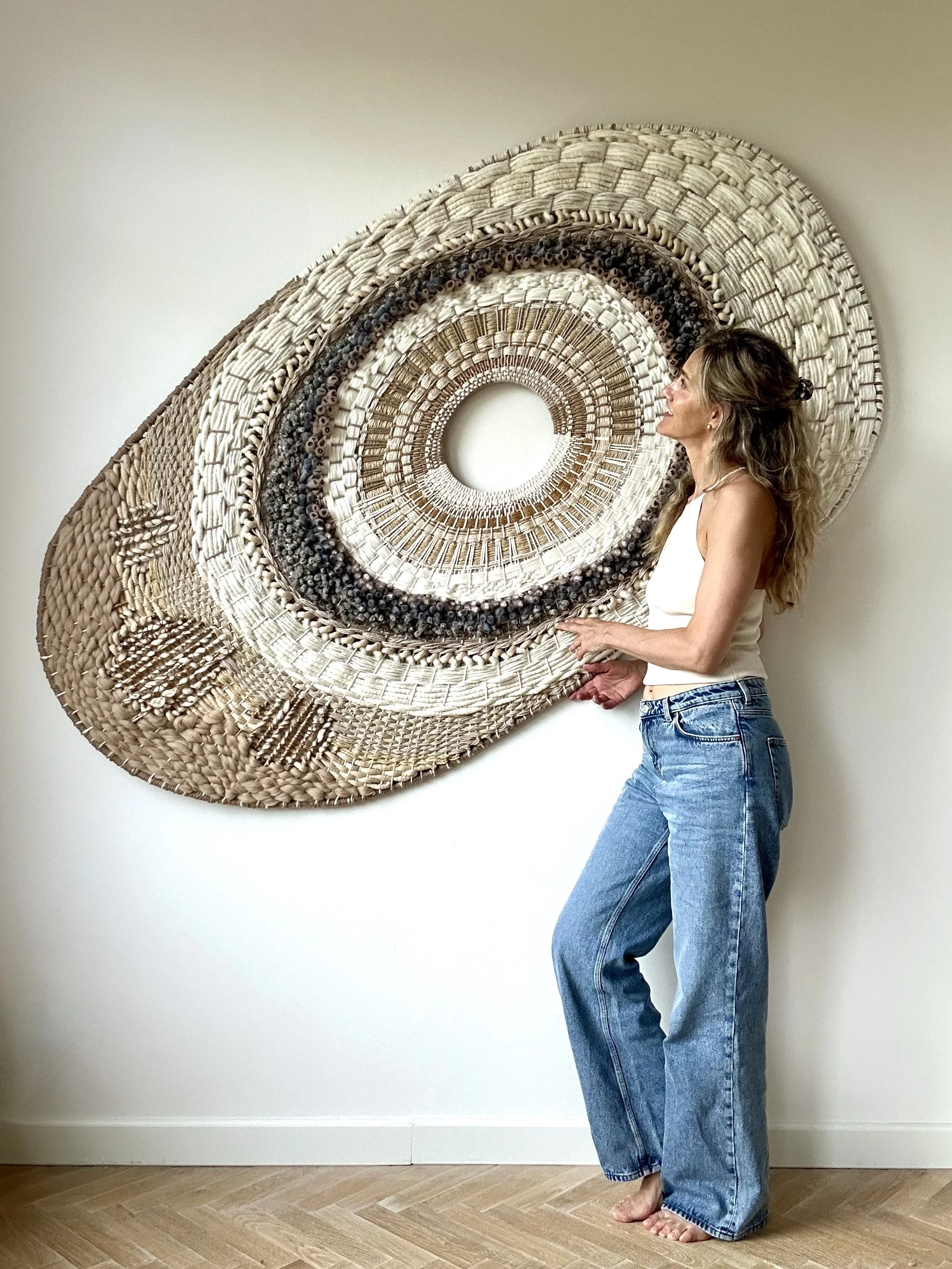 Woman standing next to a large circular woven wall art piece with intricate patterns, on a white wall, wearing jeans and a tank top.