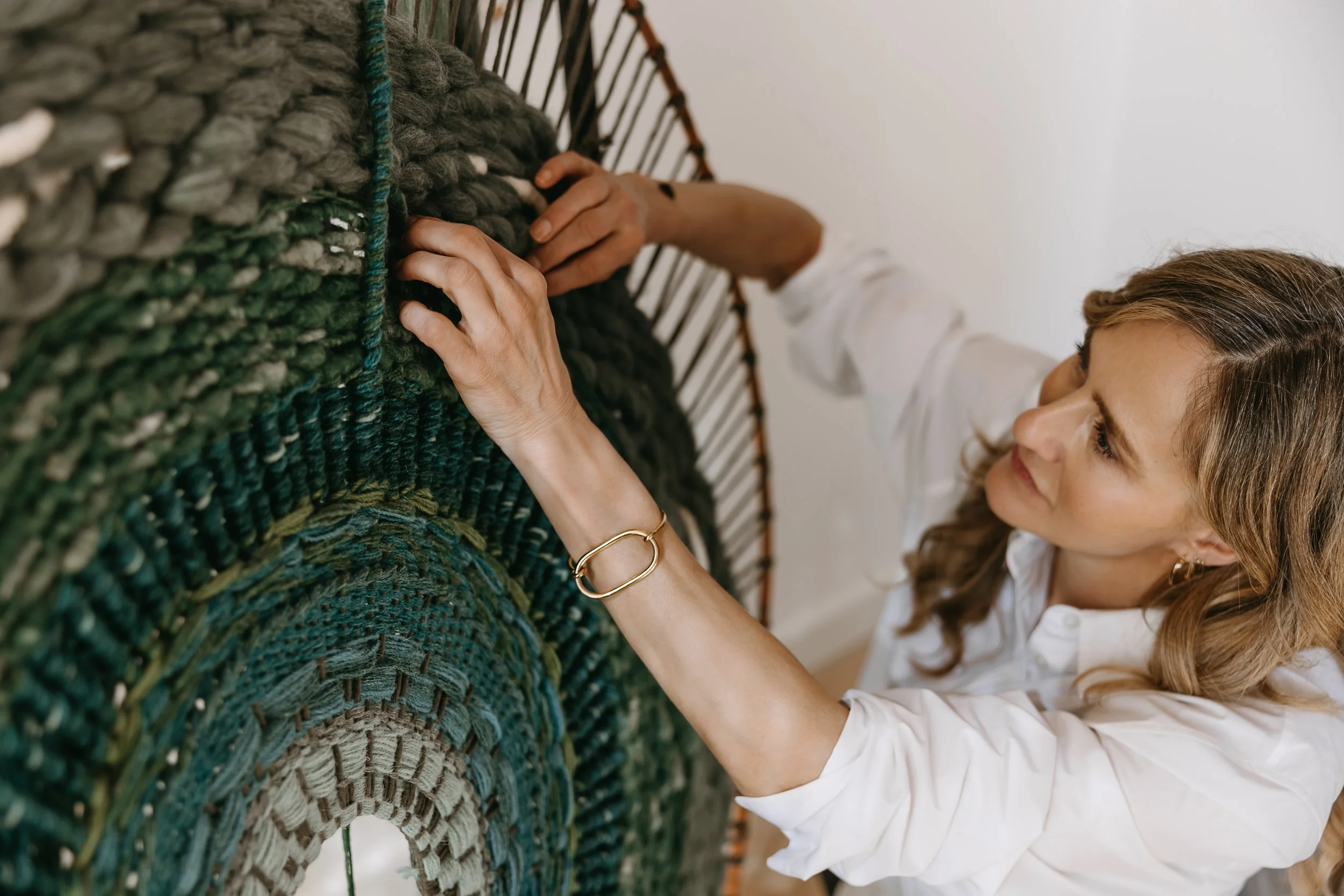 Woman weaving a large circular textile in shades of green and blue, wearing a white shirt and gold bracelet.