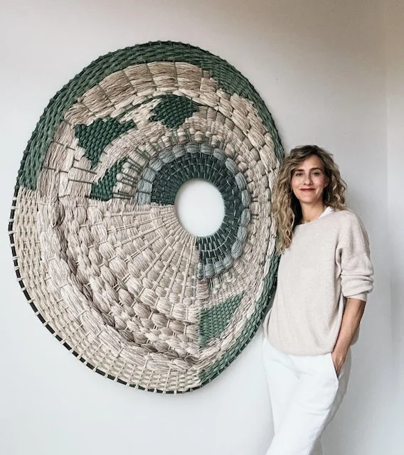 A woman standing next to a large circular woven wall art piece with a mix of green and beige tones.