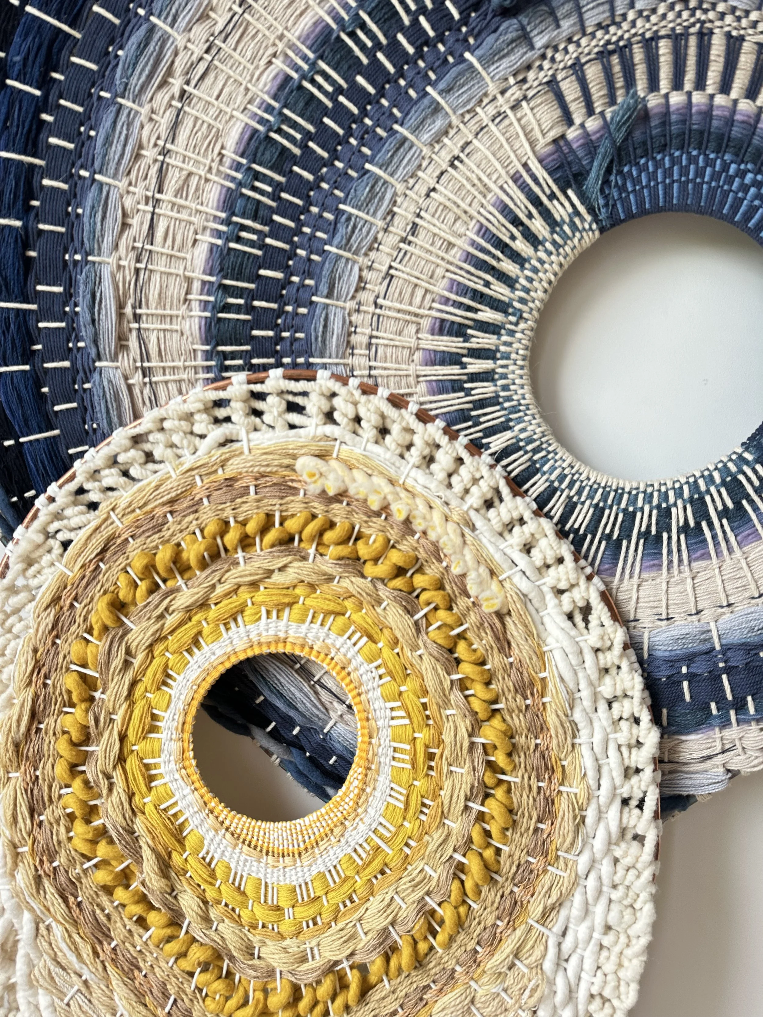 Close-up of woven circular textile art with concentric patterns in blue, beige, yellow, and brown colors.