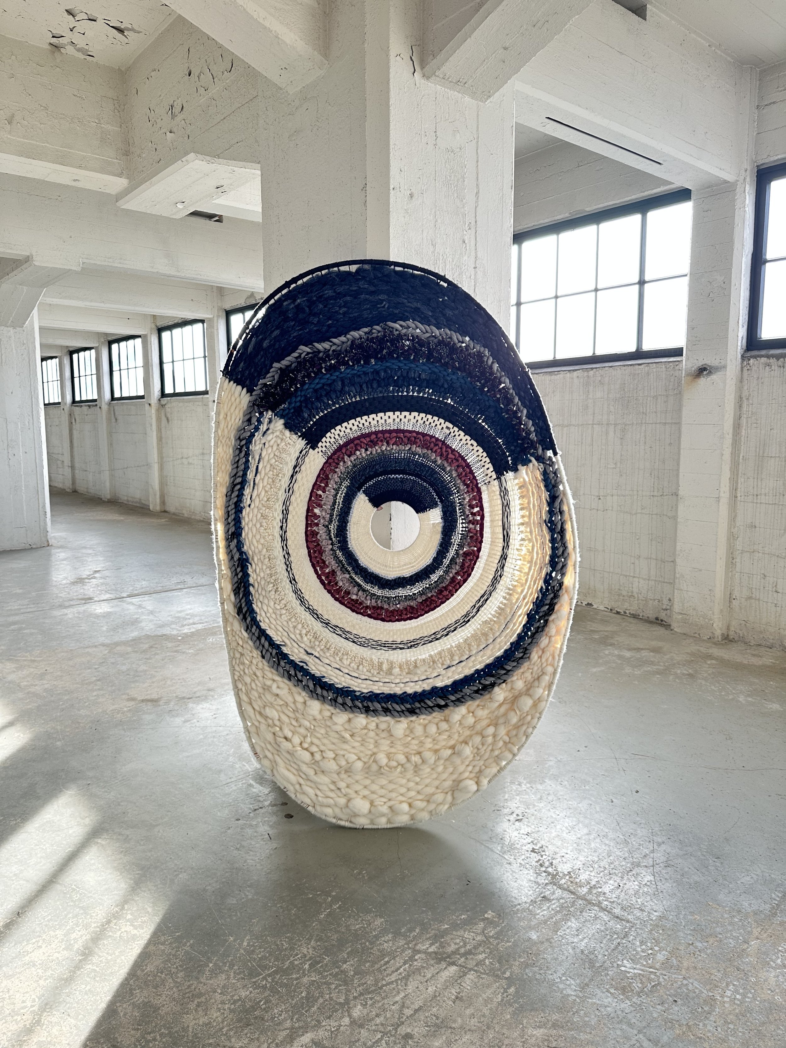 An intricate, oval-shaped woven art installation displayed in an industrial-style room, featuring concentric circles in shades of cream, navy, and burgundy.