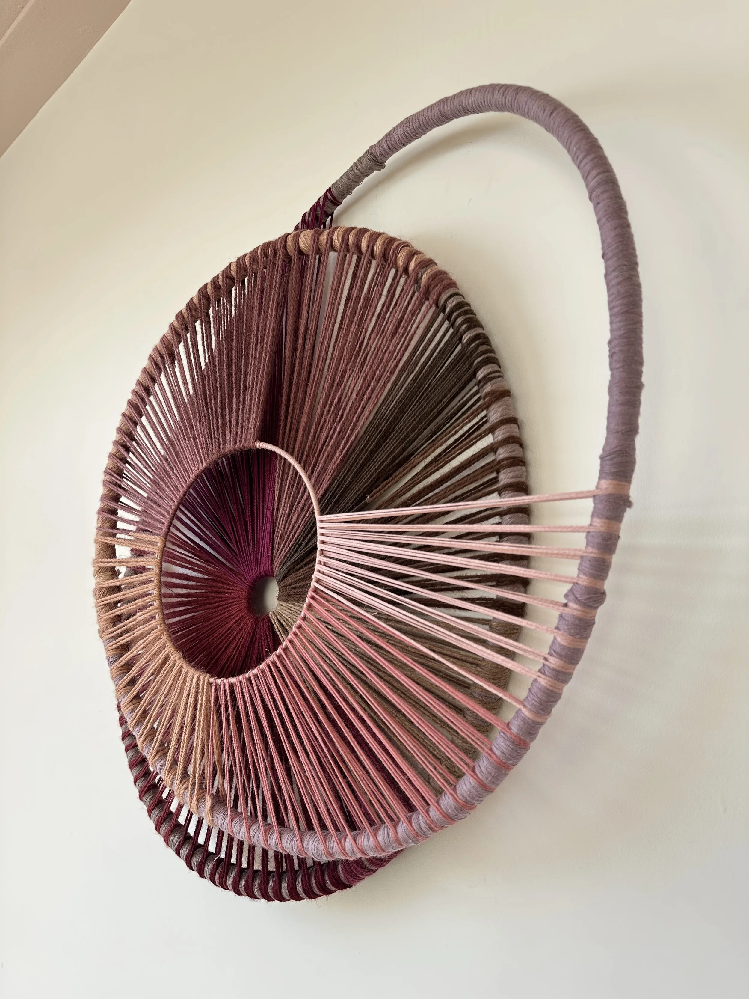 Colorful circular wall hanging made of wrapped yarn with concentric rings and earthy tones on a light background.