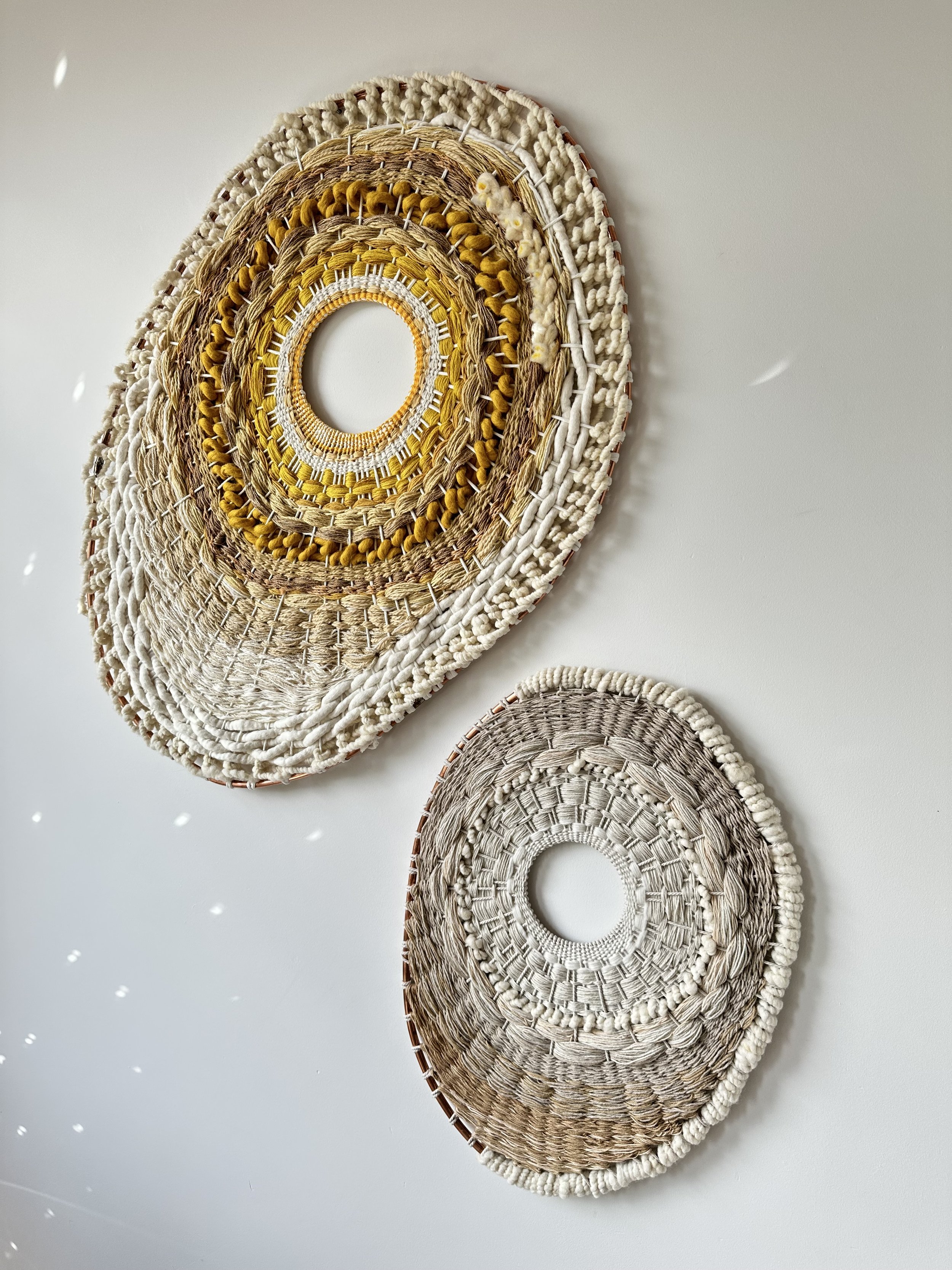 Two woven wall decorations with circular designs, one larger with earthy tones and yellow accents, and one smaller in neutral tones, hanging on a white wall with light reflections.