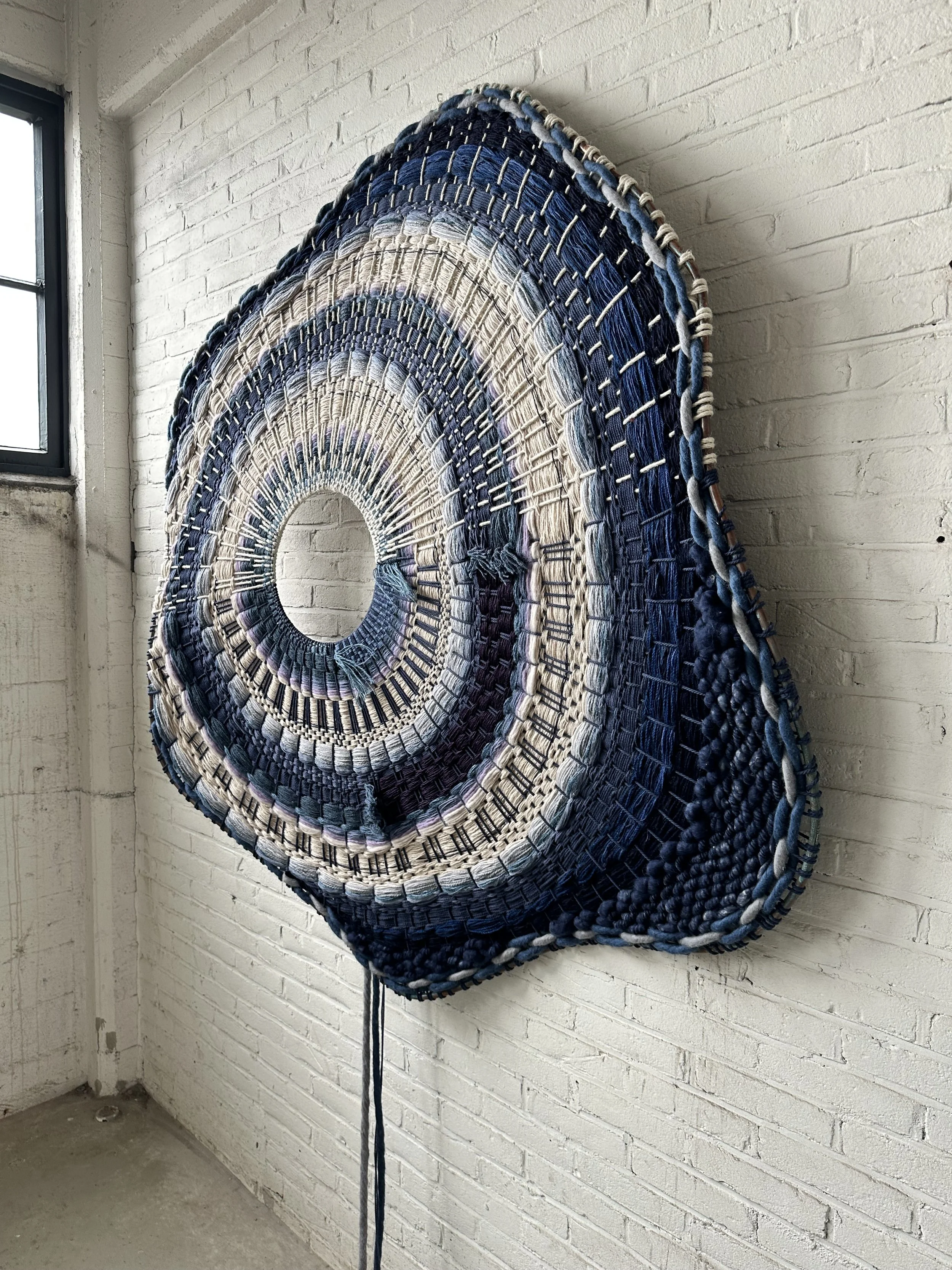 Large woven wall art with blue and beige circular patterns on a white brick wall.