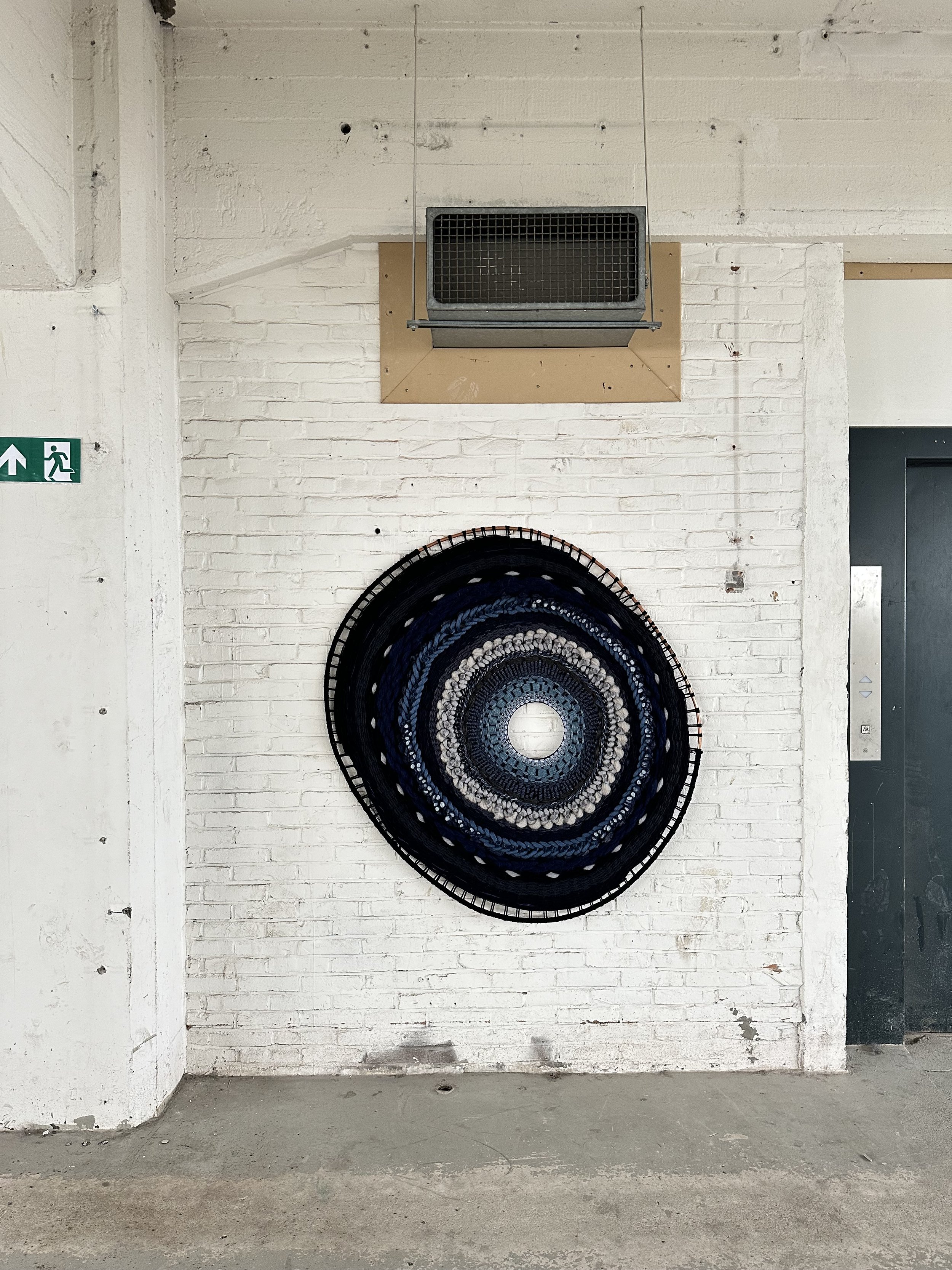 Round knitted wall art with concentric patterns in black, blue, and gray on a white brick wall near a vent and exit sign.