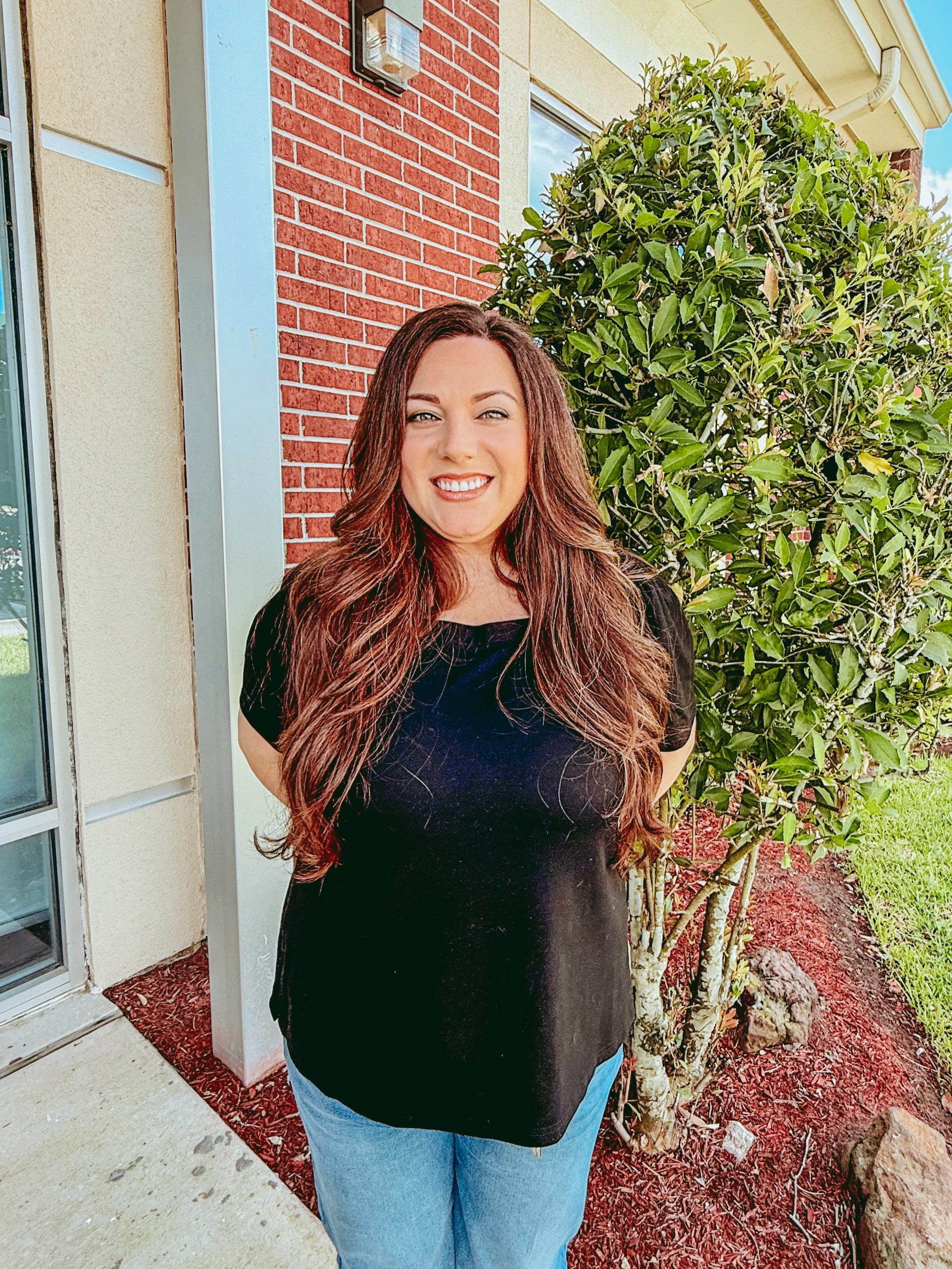 A young woman with long curly hair standing outdoors in front of a green bush, wearing a pink t-shirt and black pants, smiling at the camera.