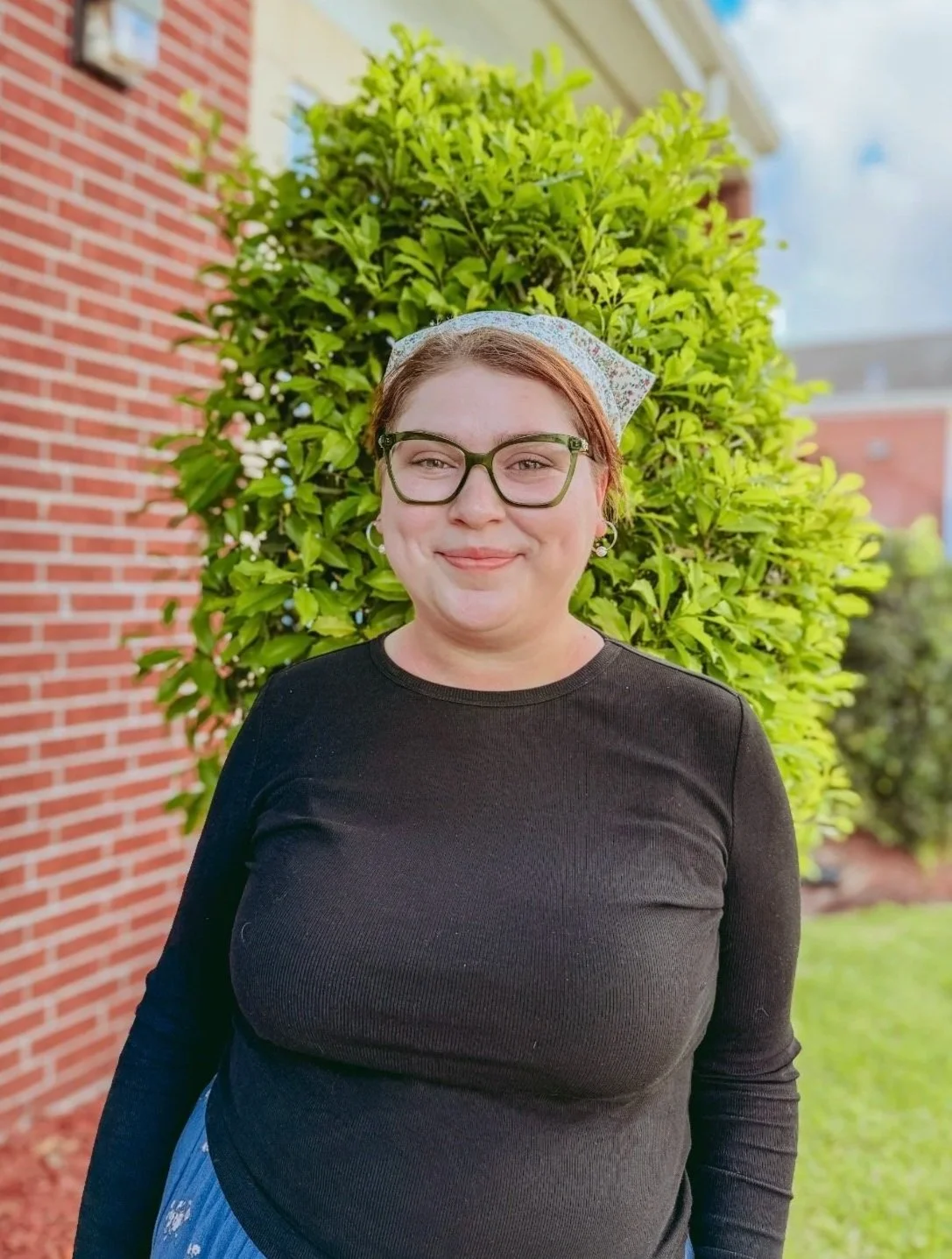 A woman with glasses and earrings smiling outdoors next to green bushes and a house window, wearing a Monument Christian Academy t-shirt.