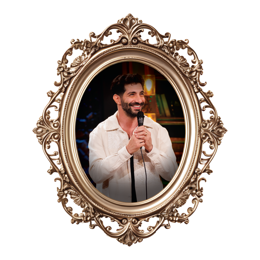 A man with dark hair and beard, smiling and holding a microphone, dressed in a white jacket, standing in front of a bookshelf and colorful background, framed by an ornate gold mirror.