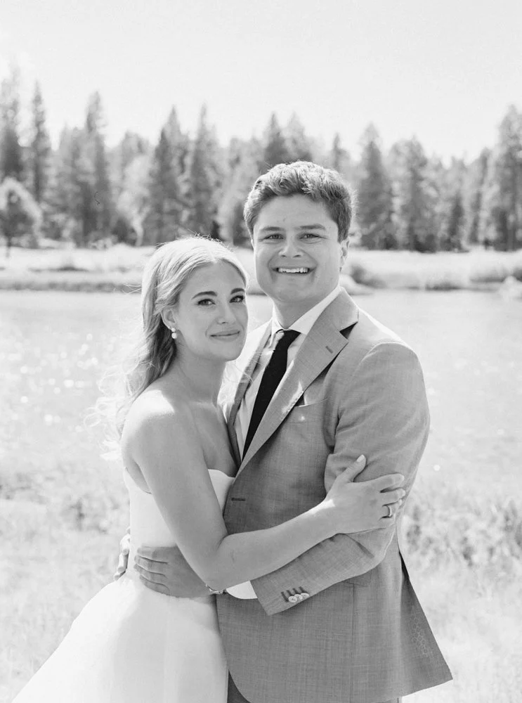A black and white photo of a couple embracing outdoors, dressed in wedding attire with a natural landscape background, including trees and a body of water.