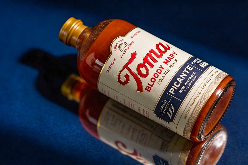A bottle of Toma Bloody Mary cocktail mixer with a reflection on a dark blue surface.